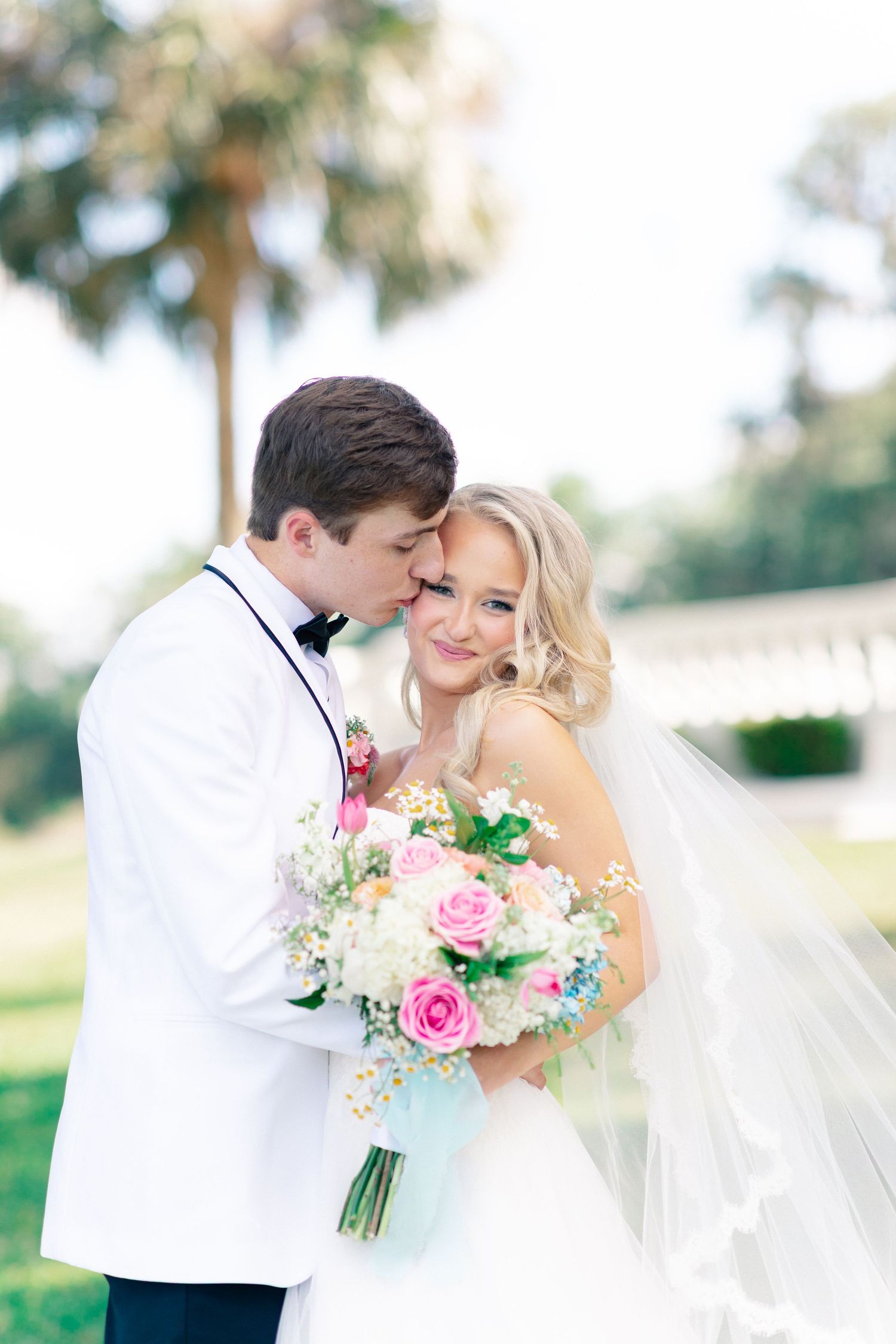 Bride and groom share a romantic embrace outdoors, bride holding a pink and white floral bouquet, groom in white tuxedo, Bella Cosa
