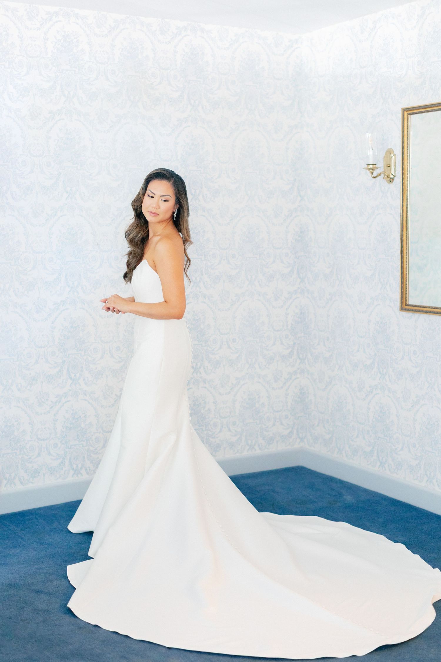 Elegant bride in a white strapless mermaid wedding gown with a long train, posing in a blue damask wallpaper room.