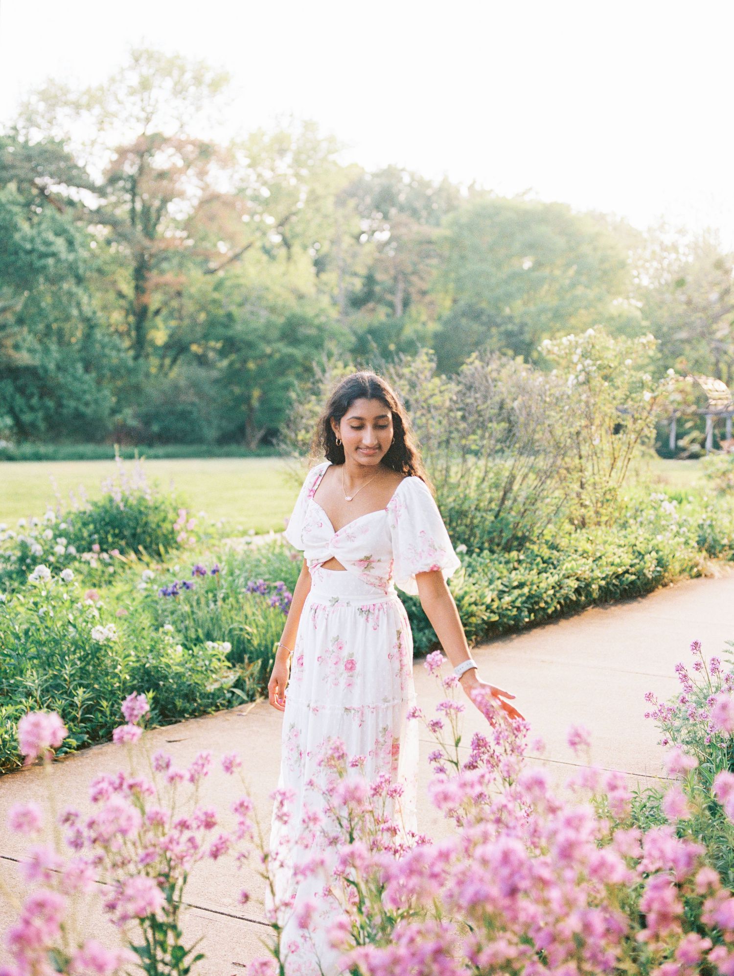 Woman in floral dress standing among pink blooms in a sunlit garden path.