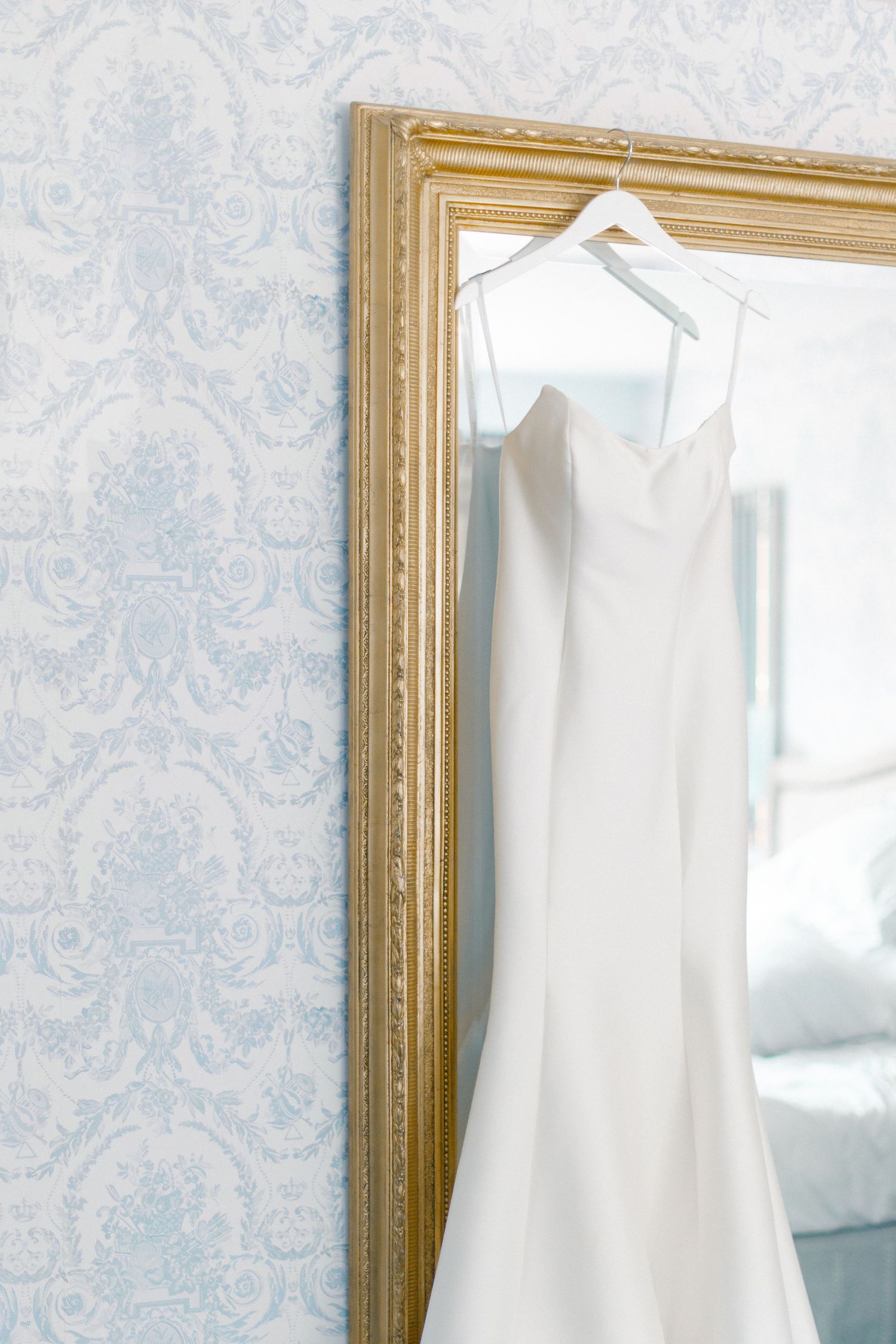 Elegant white wedding gown hanging on a gold-framed mirror against blue damask wallpaper.