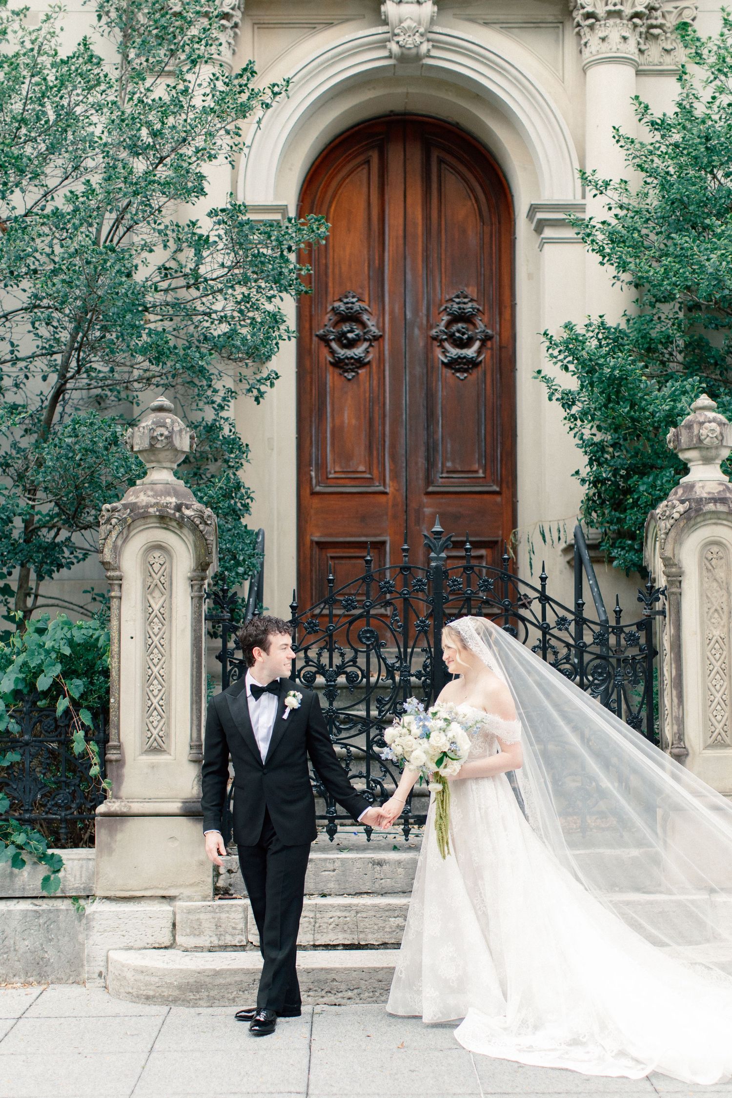 Bride and groom hold hands before ornate wooden door with iron gate, her veil flowing, he in tuxedo.