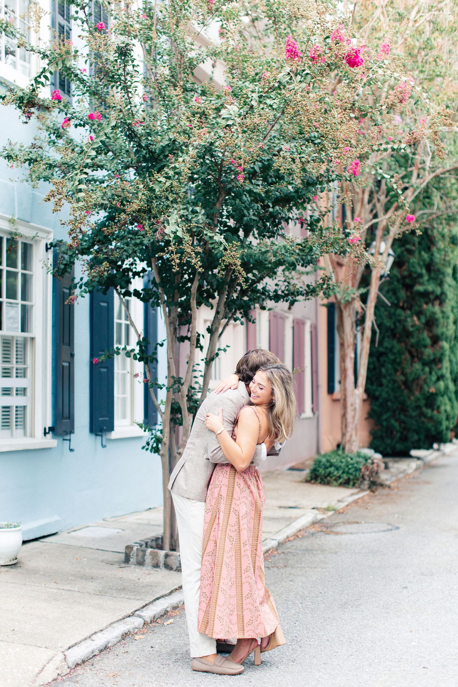 Couple sharing a romantic hug on a colorful Charleston street lined with pastel historic buildings and blooming trees.