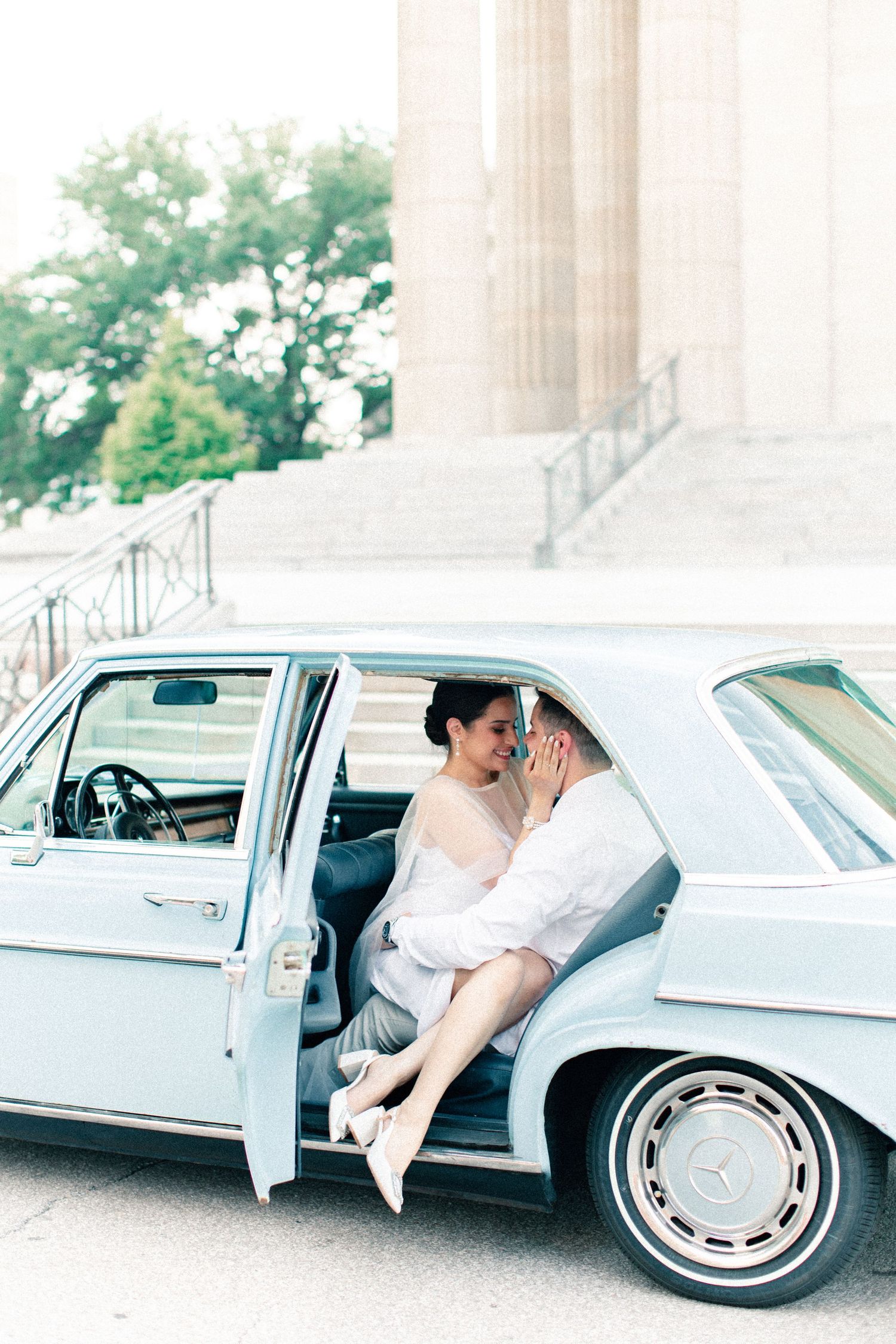 Bride and groom sitting in a vintage light blue Mercedes car outside a classical building during their wedding day.