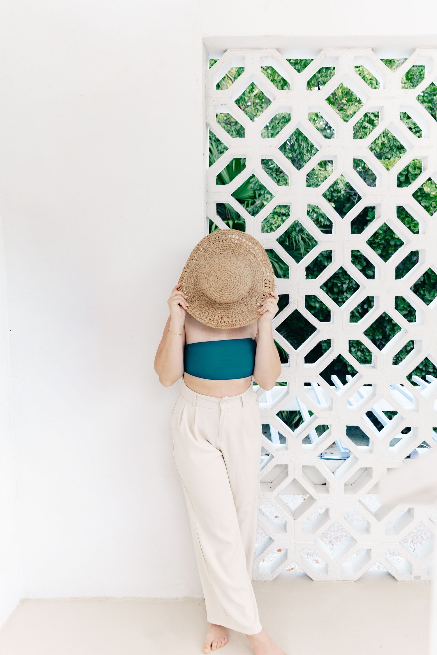 Woman in teal crop top, cream pants, and straw hat standing by decorative white lattice wall with green foliage.