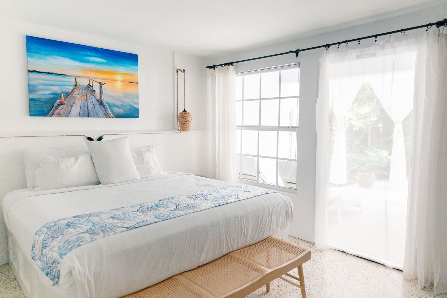 Bright white bedroom with king bed, blue accent runner, coastal sunset artwork, and French doors with sheer curtains.
