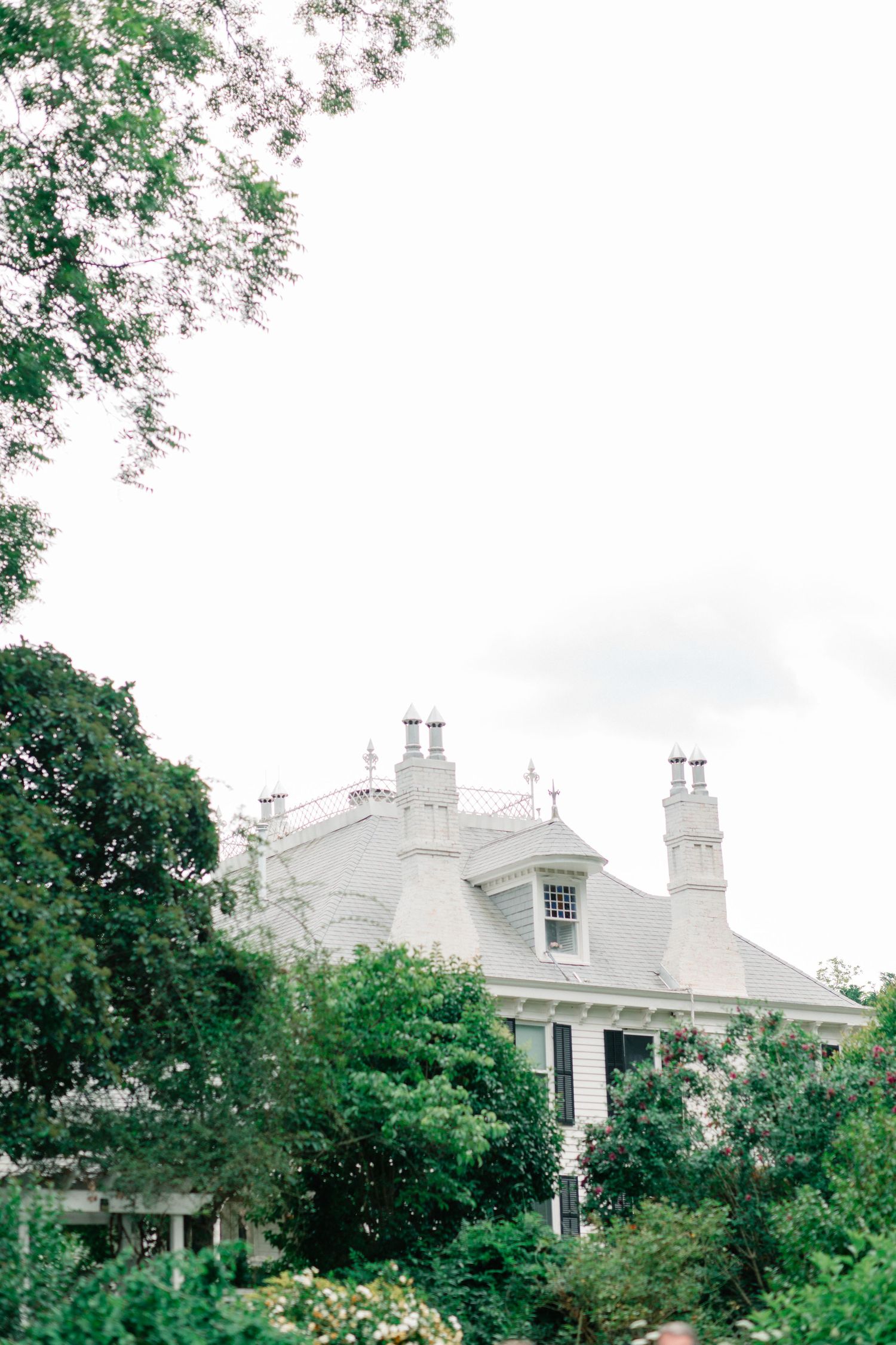 Elegant white mansion partially obscured by lush green trees under an overcast sky.