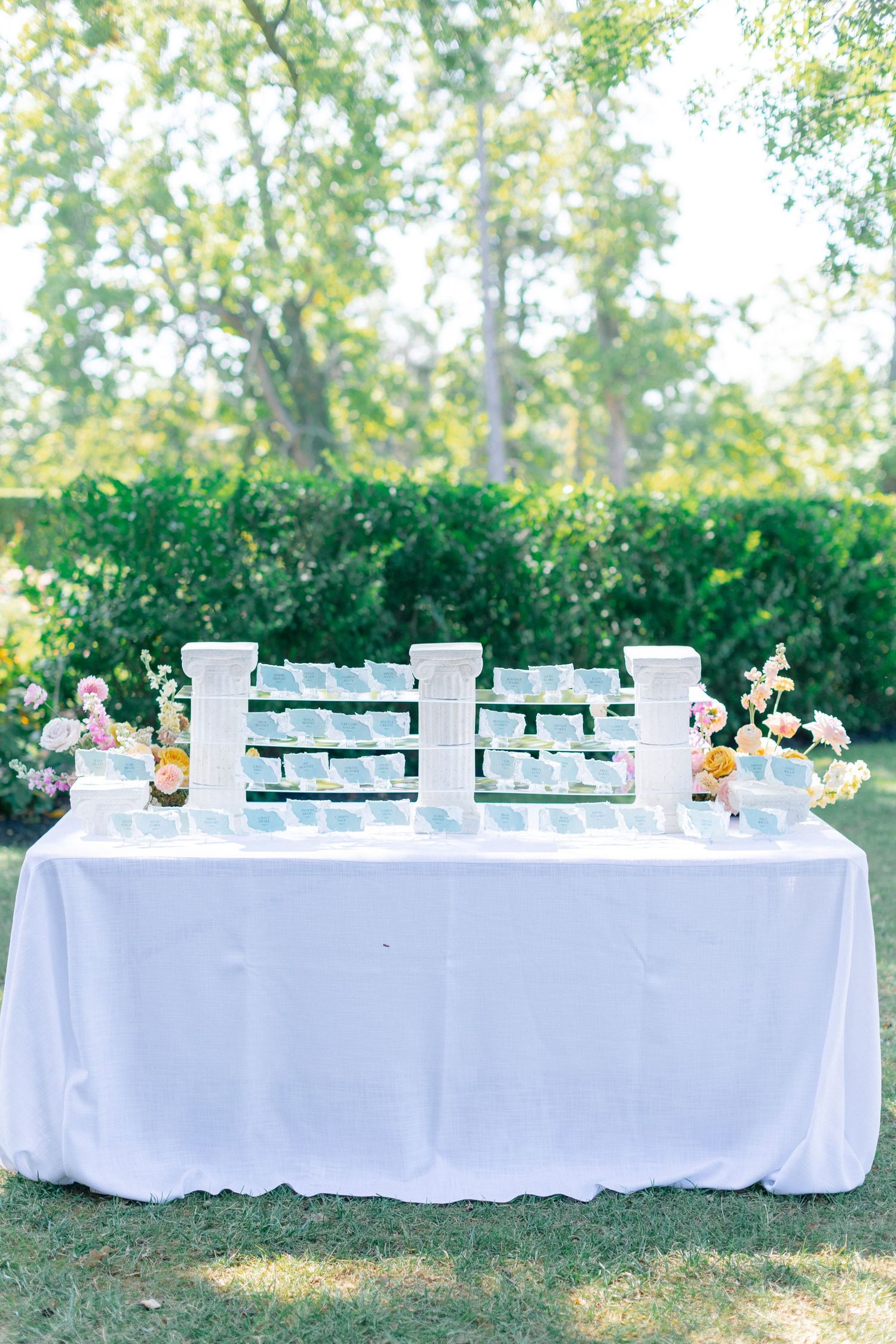 Elegant outdoor wedding escort card table with white tablecloth, tiered card display, and fresh floral arrangements.