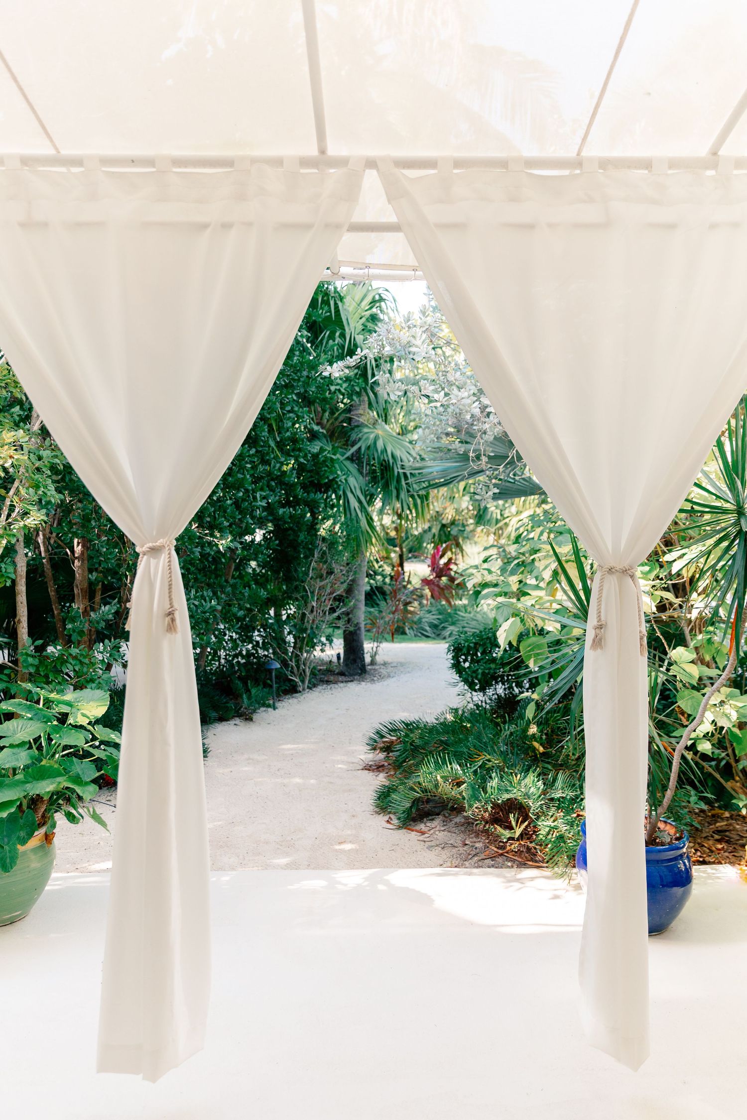 White curtained cabana opens to a lush tropical garden pathway with palm trees and blue ceramic pots.