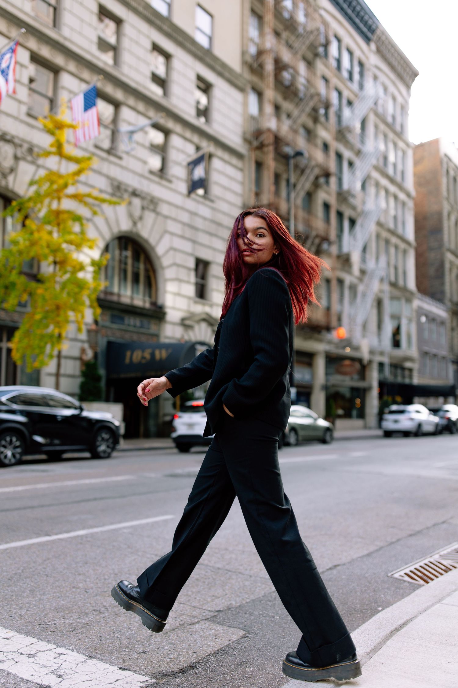 Young woman with red hair in all-black outfit crossing a city street, with urban buildings in the background.