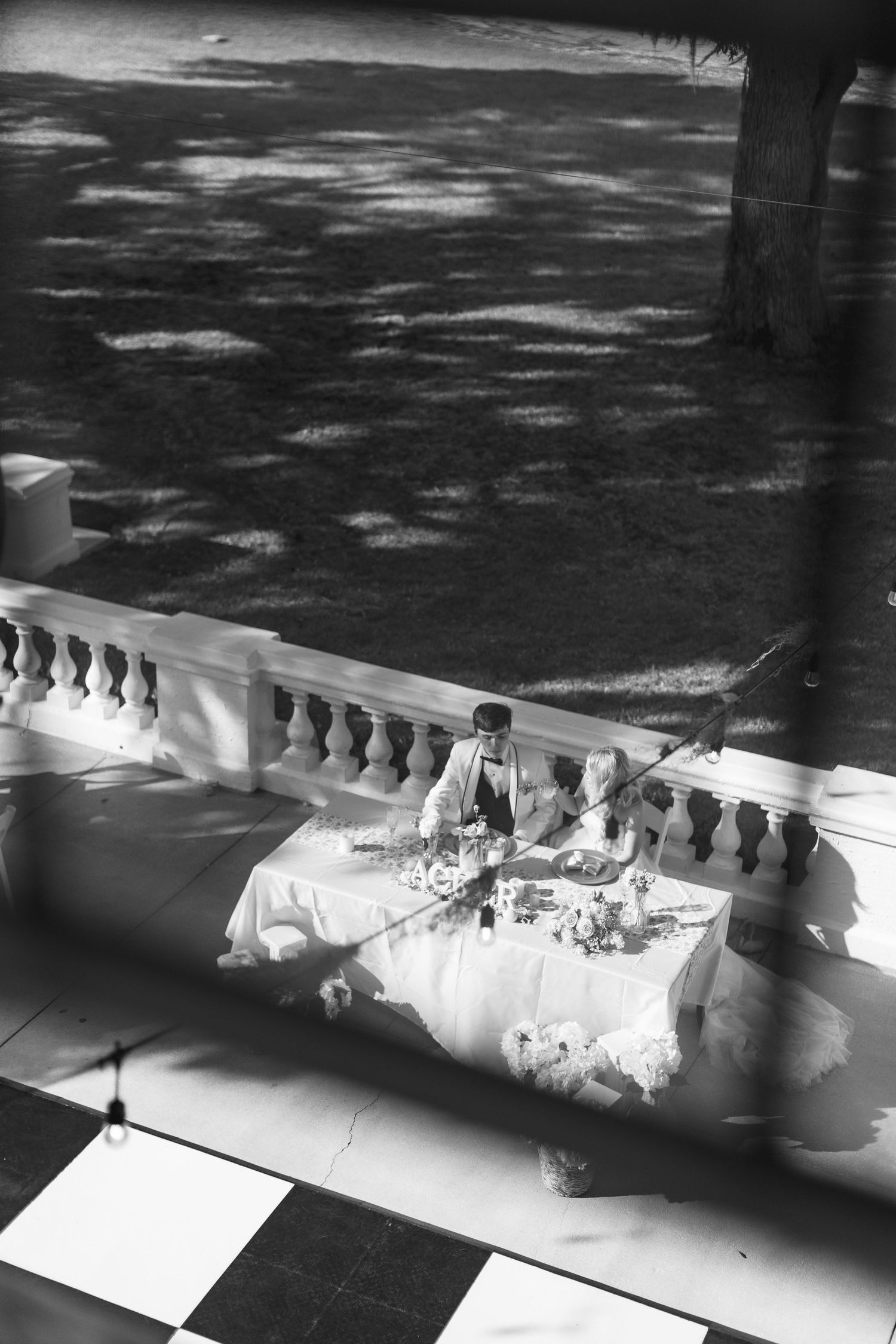 Black and white aerial view of a wedding couple seated at an elegant table on a balcony with ornate railings.