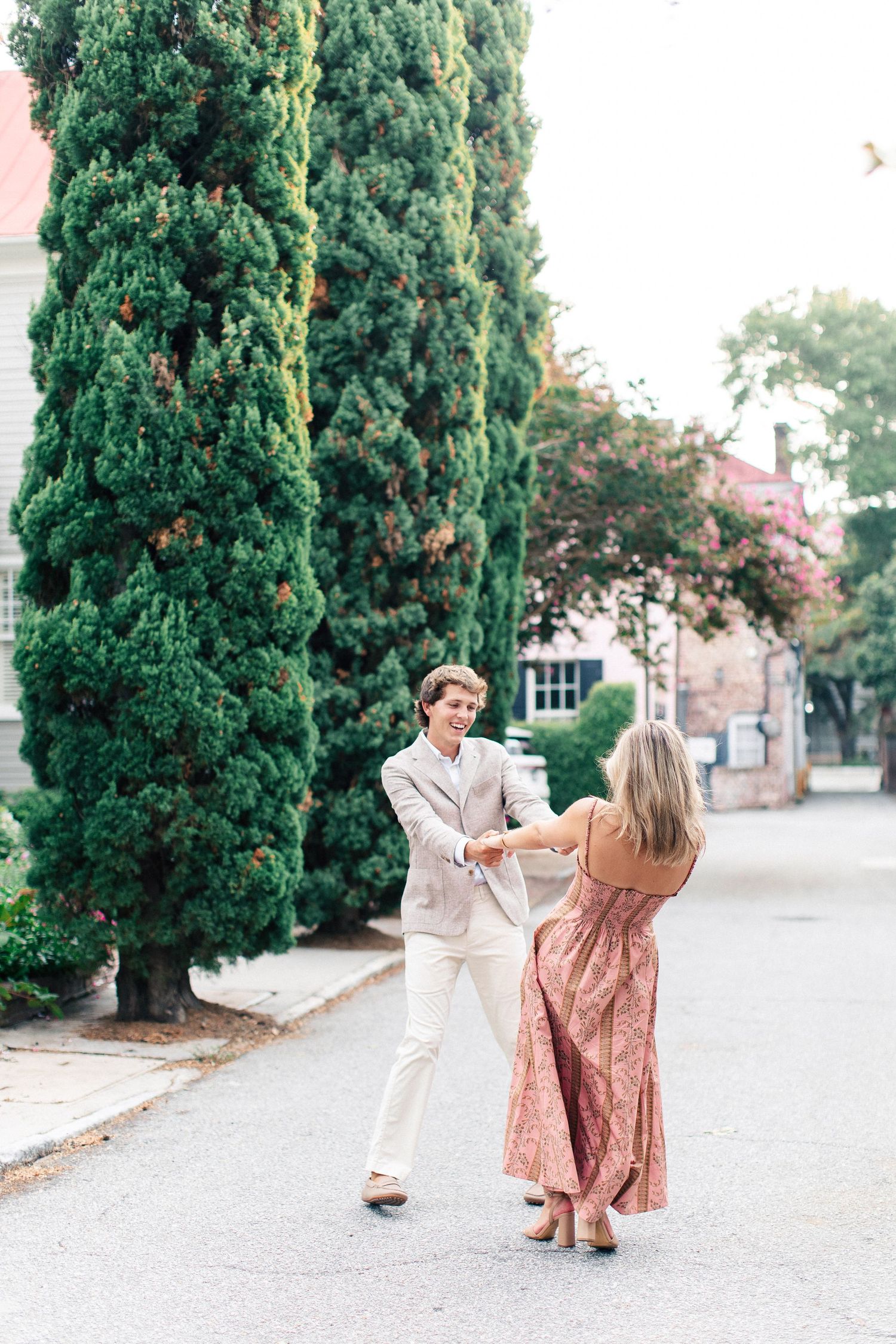 A joyful couple dances together on a street beside tall cypress trees during their engagement shoot.
