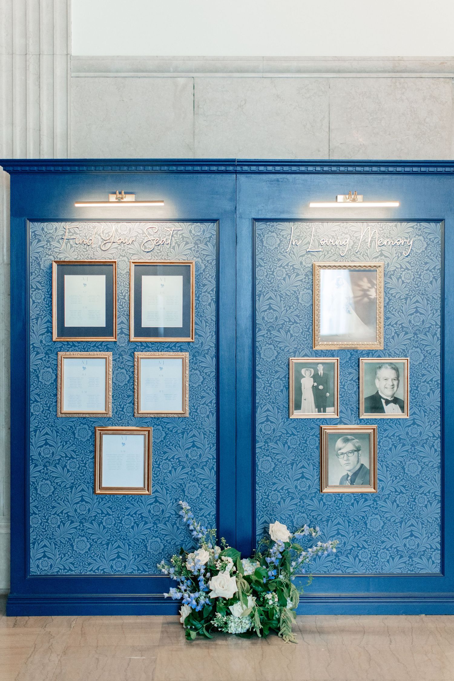 Blue decorative wall display with gold-framed photos and floral arrangement, memorial tribute with elegant wallpaper backdrop.