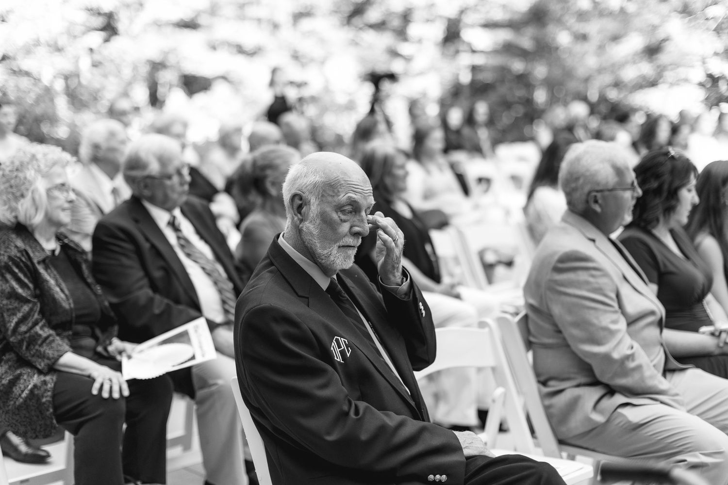 Emotional elderly wedding guest wiping tears during outdoor ceremony seated among other guests in black and white photo.