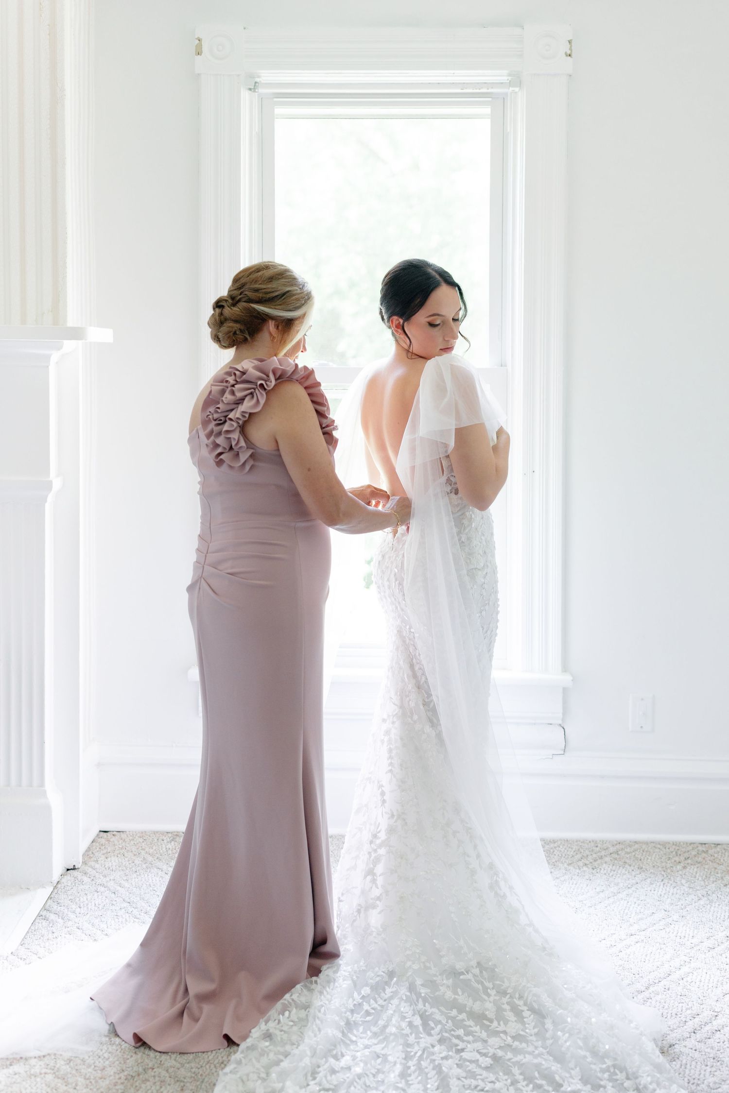 Bride in lace wedding gown being helped by bridesmaid in mauve dress near bright window during wedding preparations.