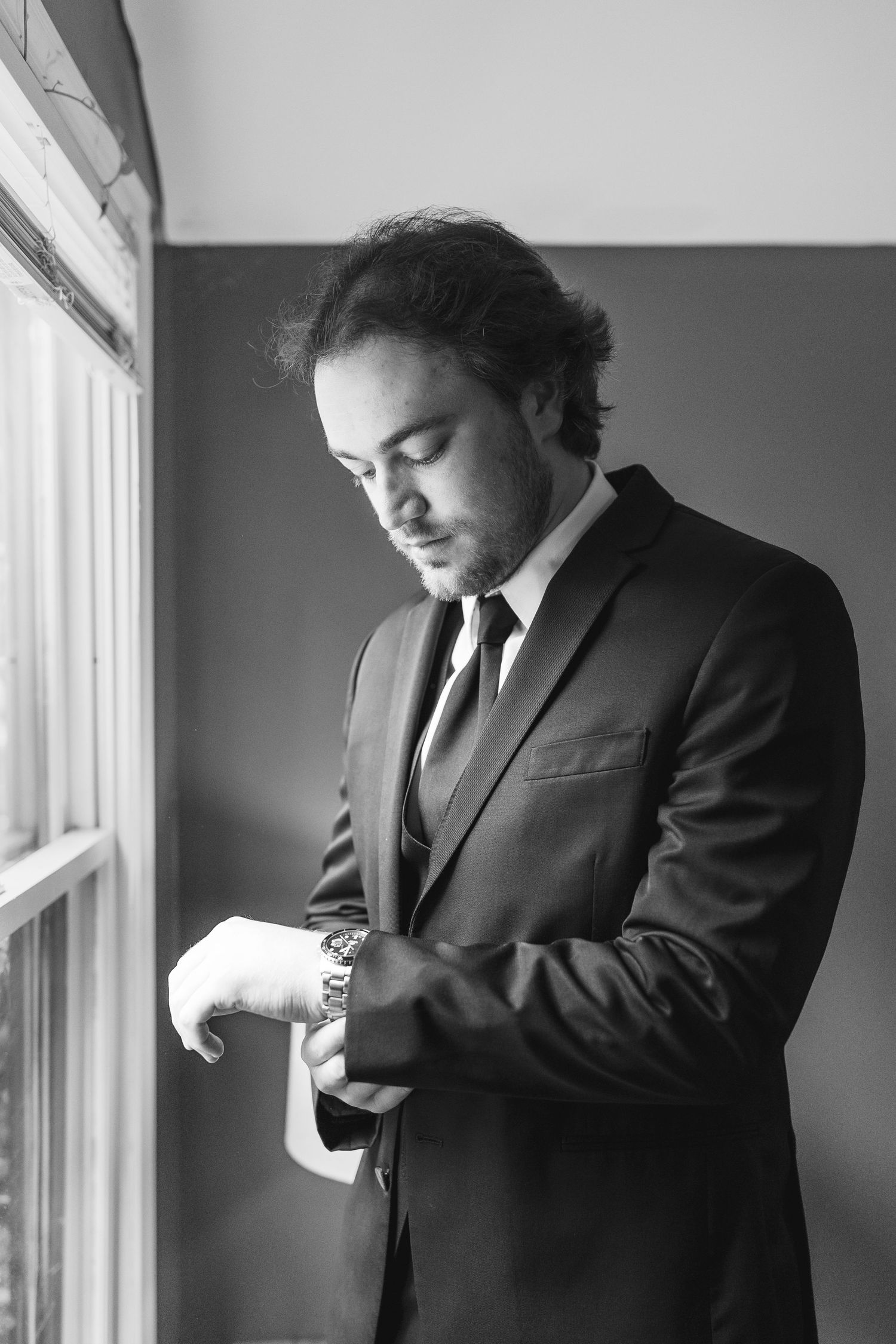 Black and white photo of a well-dressed man in a suit adjusting his watch cufflinks by a window.
