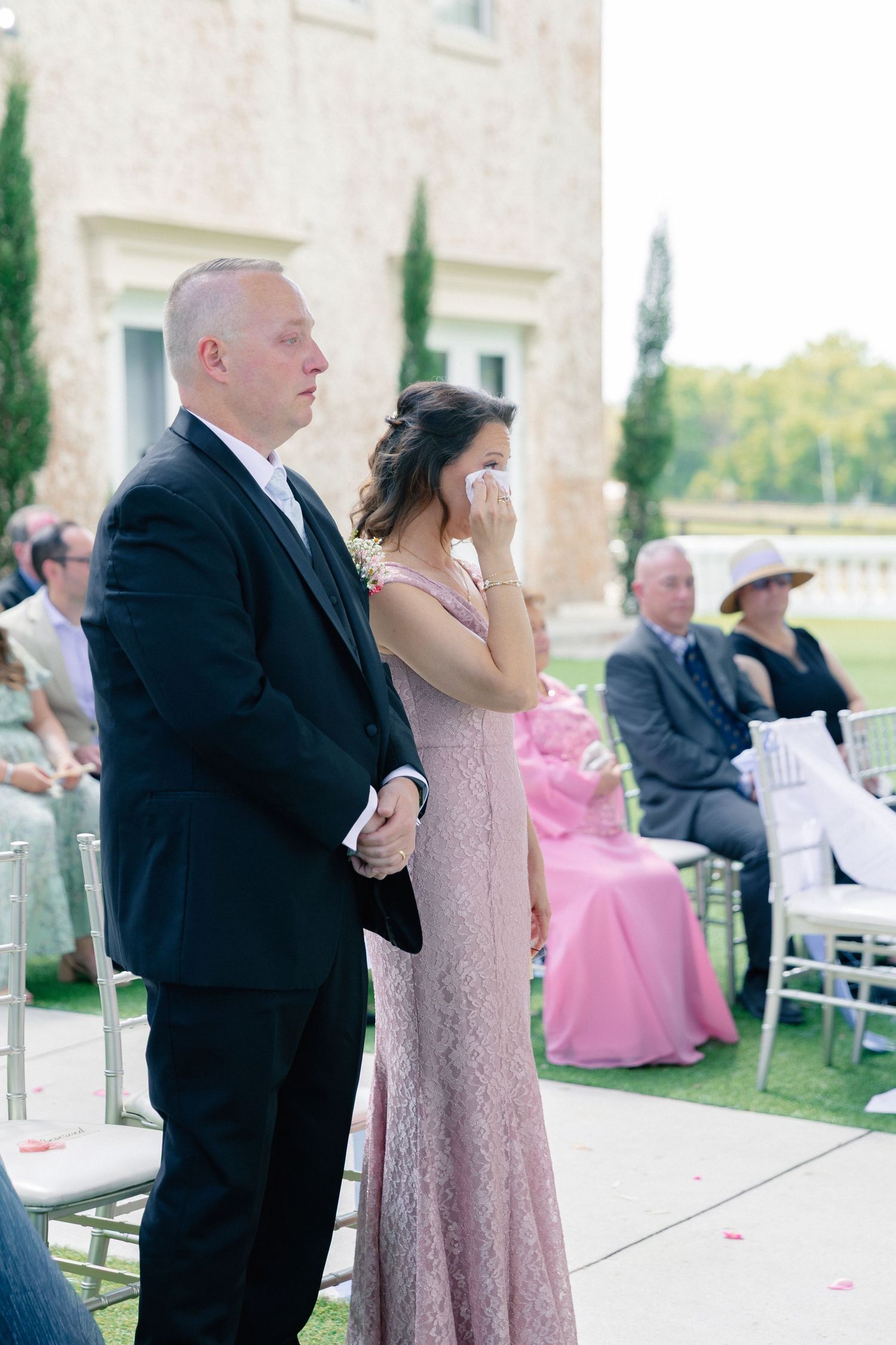 A man in a black suit and woman in pink lace dress stand emotional at an outdoor wedding ceremony, wedding at Bella Cosa
