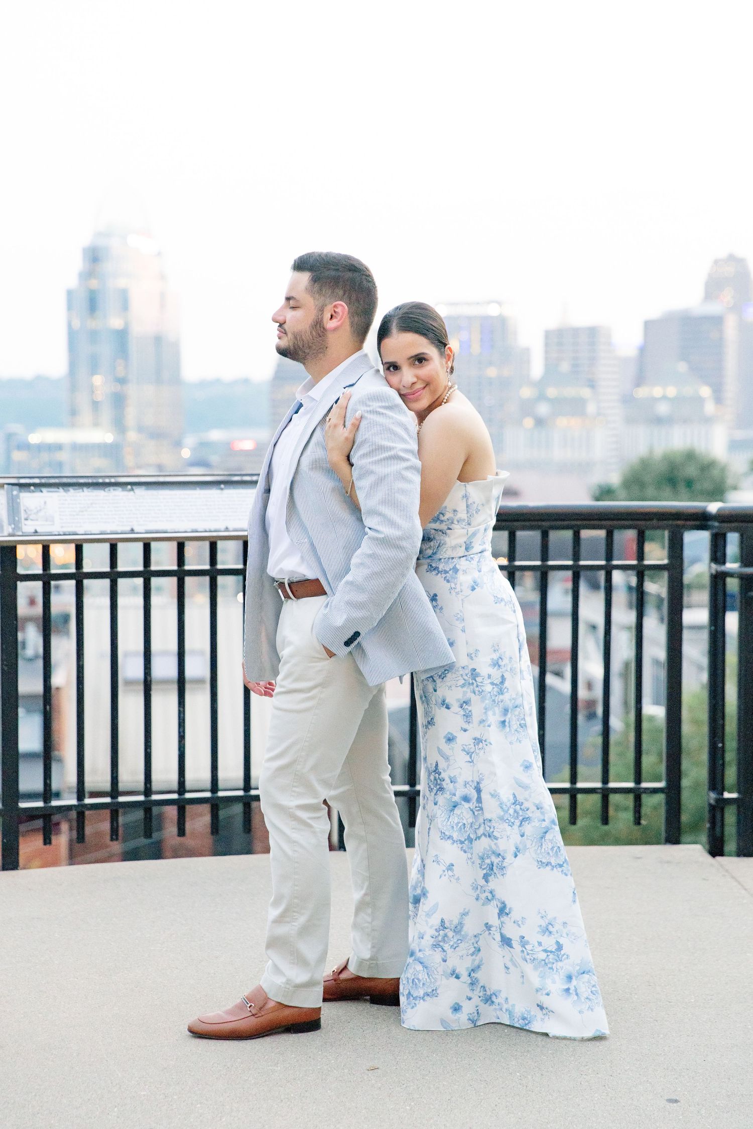 Bride in a blue floral gown and groom in a light suit stand back to back smiling on a city rooftop.