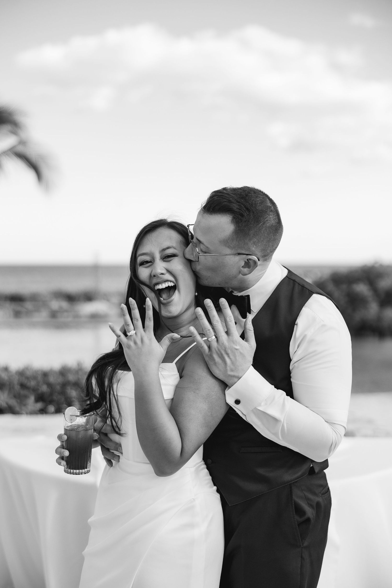 Black and white photo of a joyful bride and groom showing wedding rings, sharing a kiss outdoors near the ocean.