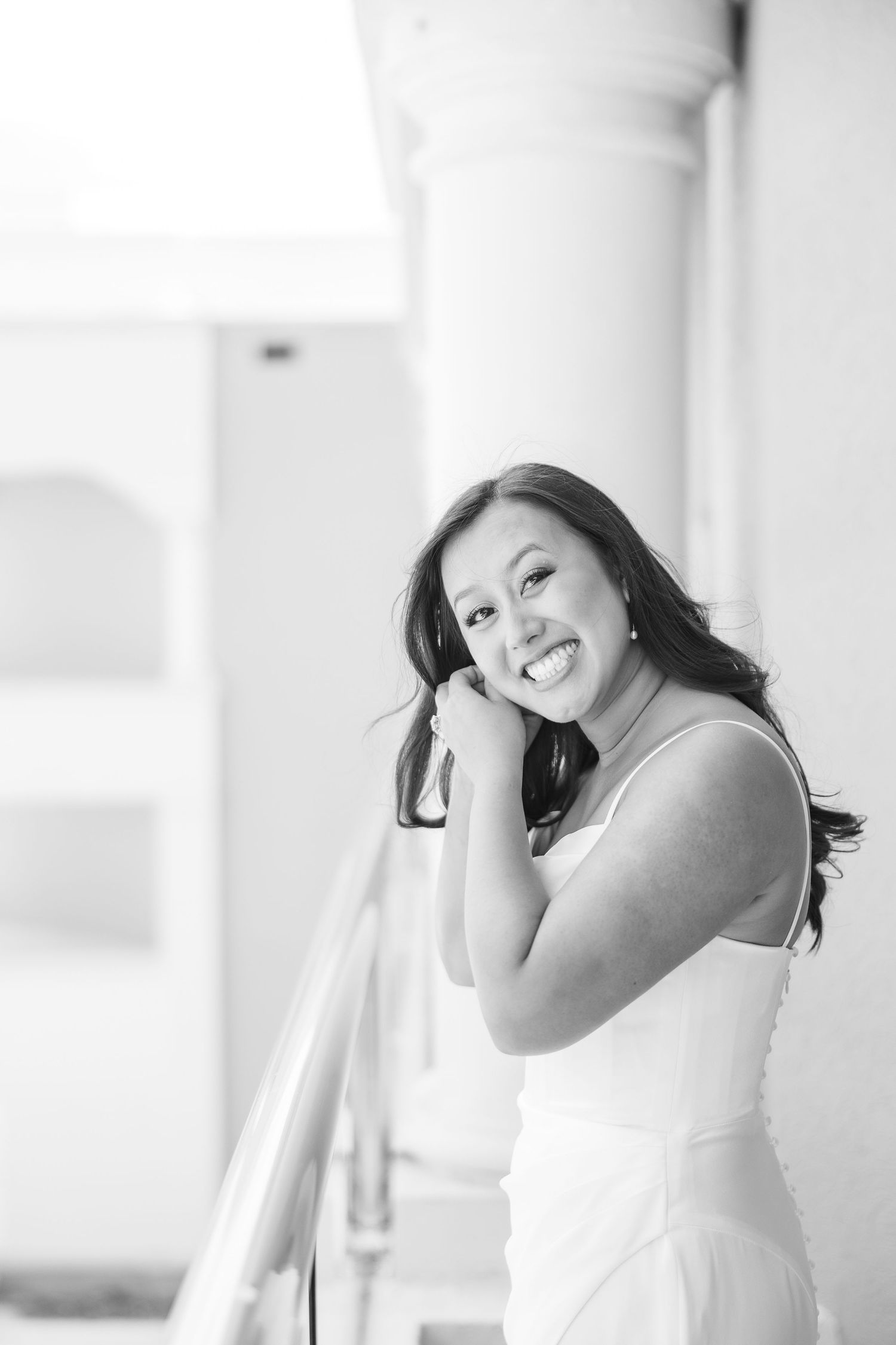 Smiling young woman in white dress leaning on railing, black and white portrait photography.