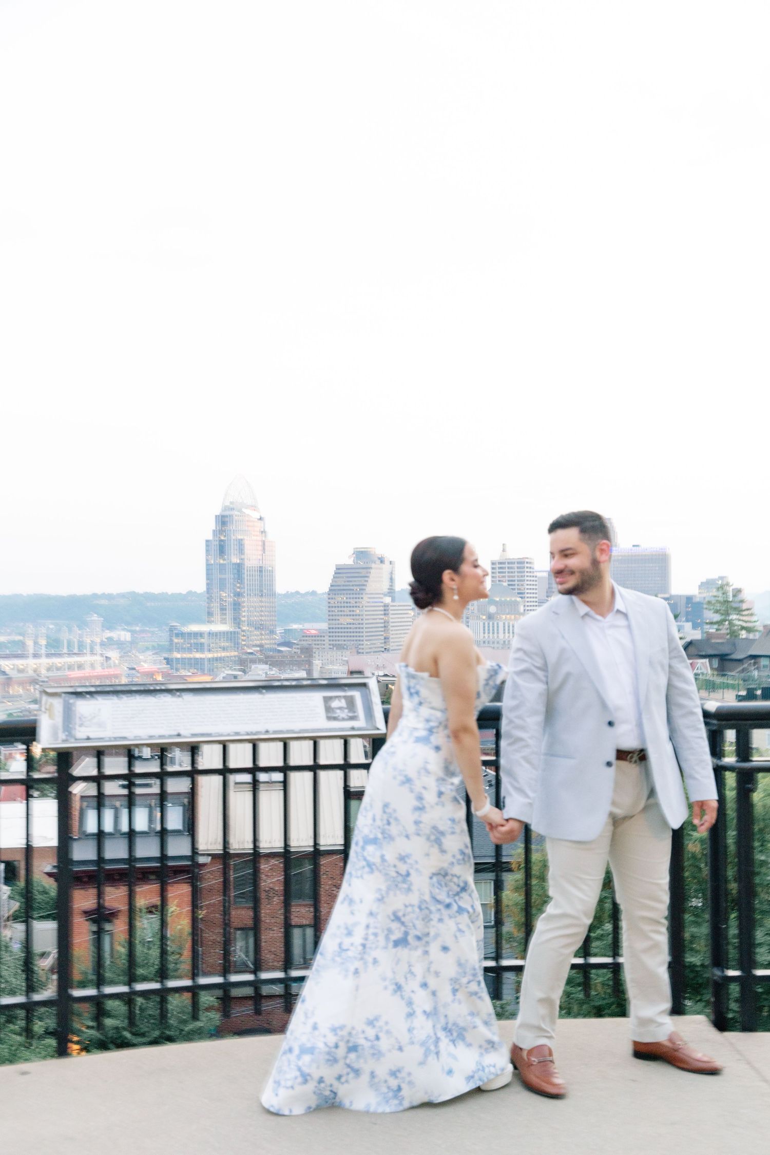Couple in coordinating blue floral and linen outfits hold hands smiling on a rooftop with city skyline behind them.