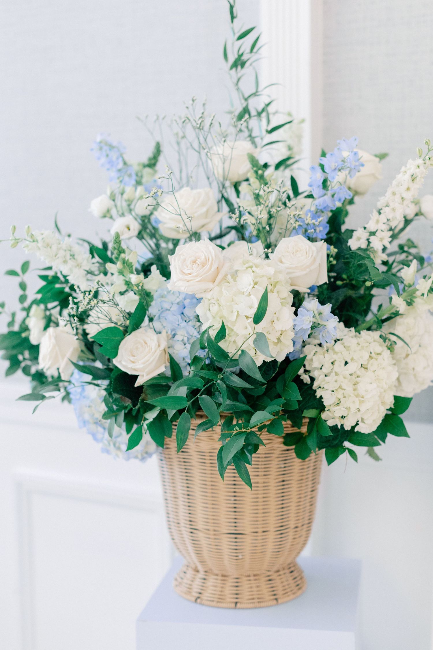 Elegant wicker basket filled with white roses, blue hydrangeas, and lush greenery arrangement.