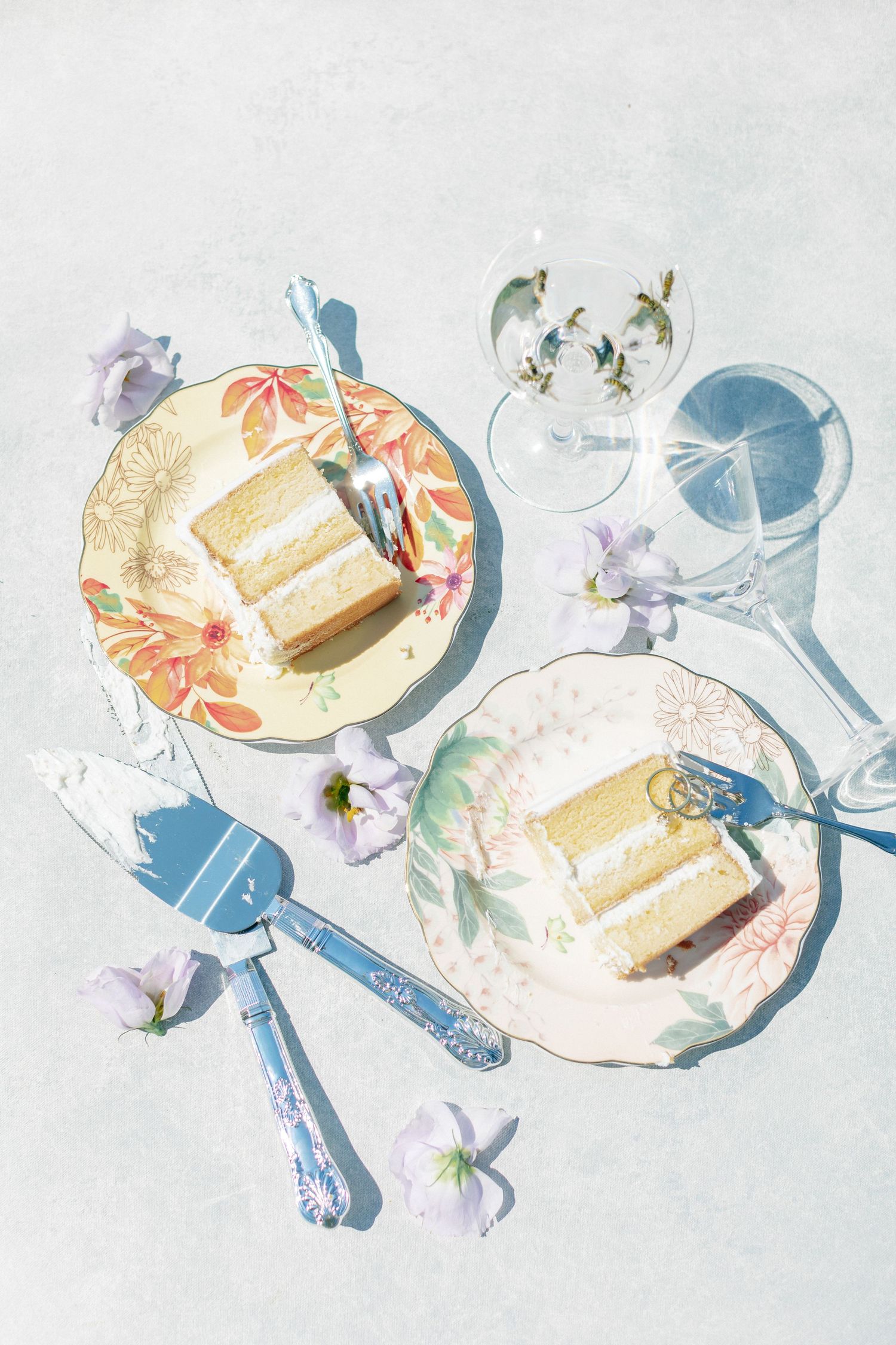 Two slices of white wedding cake on floral plates with blue cake server and champagne glasses on white surface.