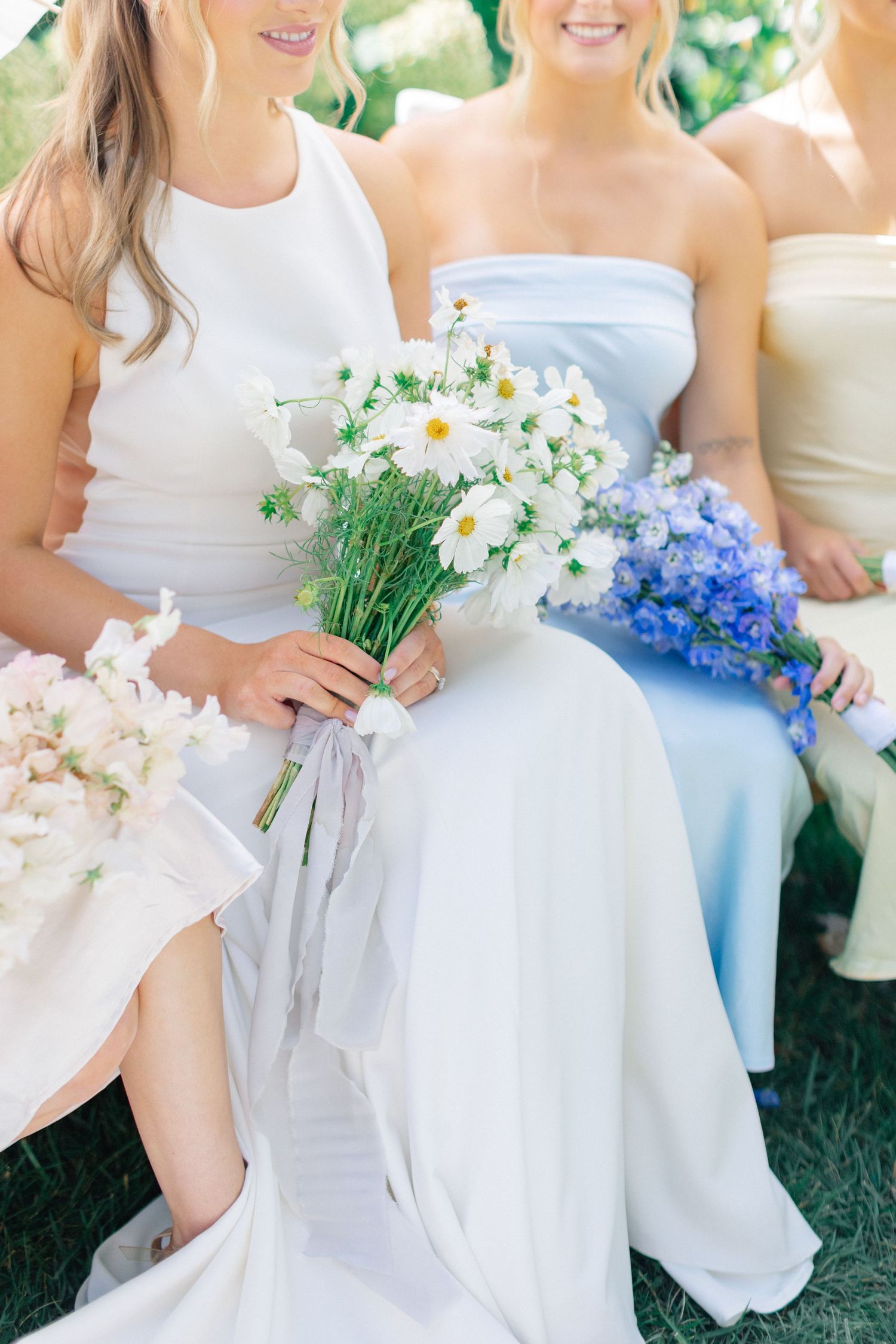 Bridesmaids in pastel gowns sitting outdoors holding bouquets of white daisies and blue flowers at a wedding.