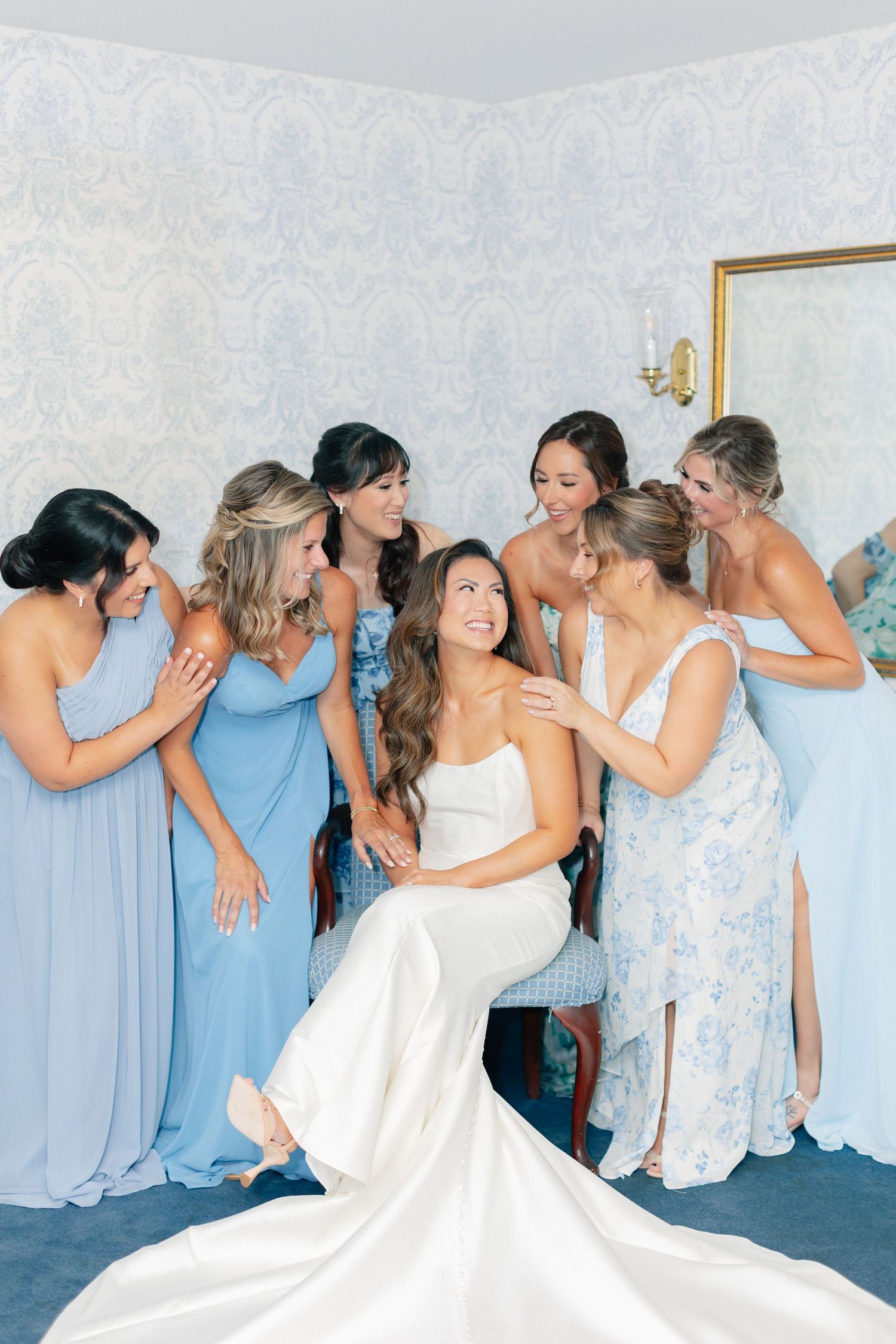 A bride in a white gown sits surrounded by bridesmaids in blue dresses during wedding preparations.