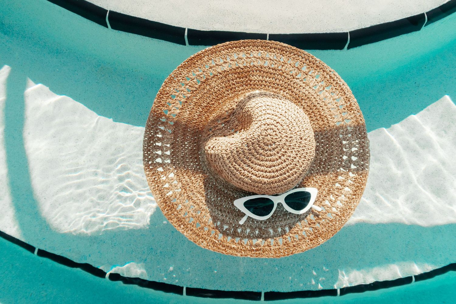 Straw sun hat and white sunglasses resting on a teal inflatable pool float in clear blue water.