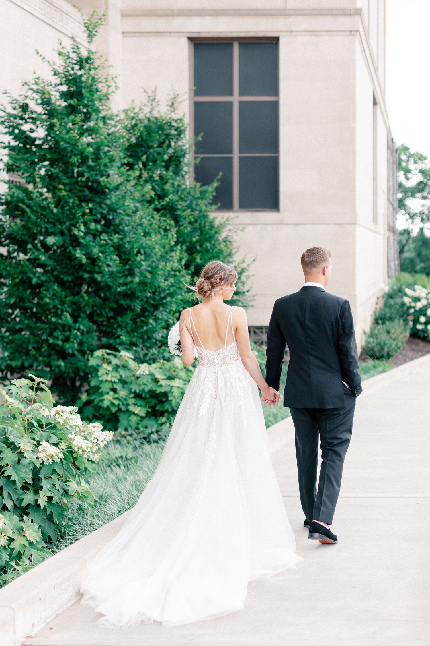 Bride and groom walk hand in hand along a garden path, her lace gown trailing elegantly behind them.