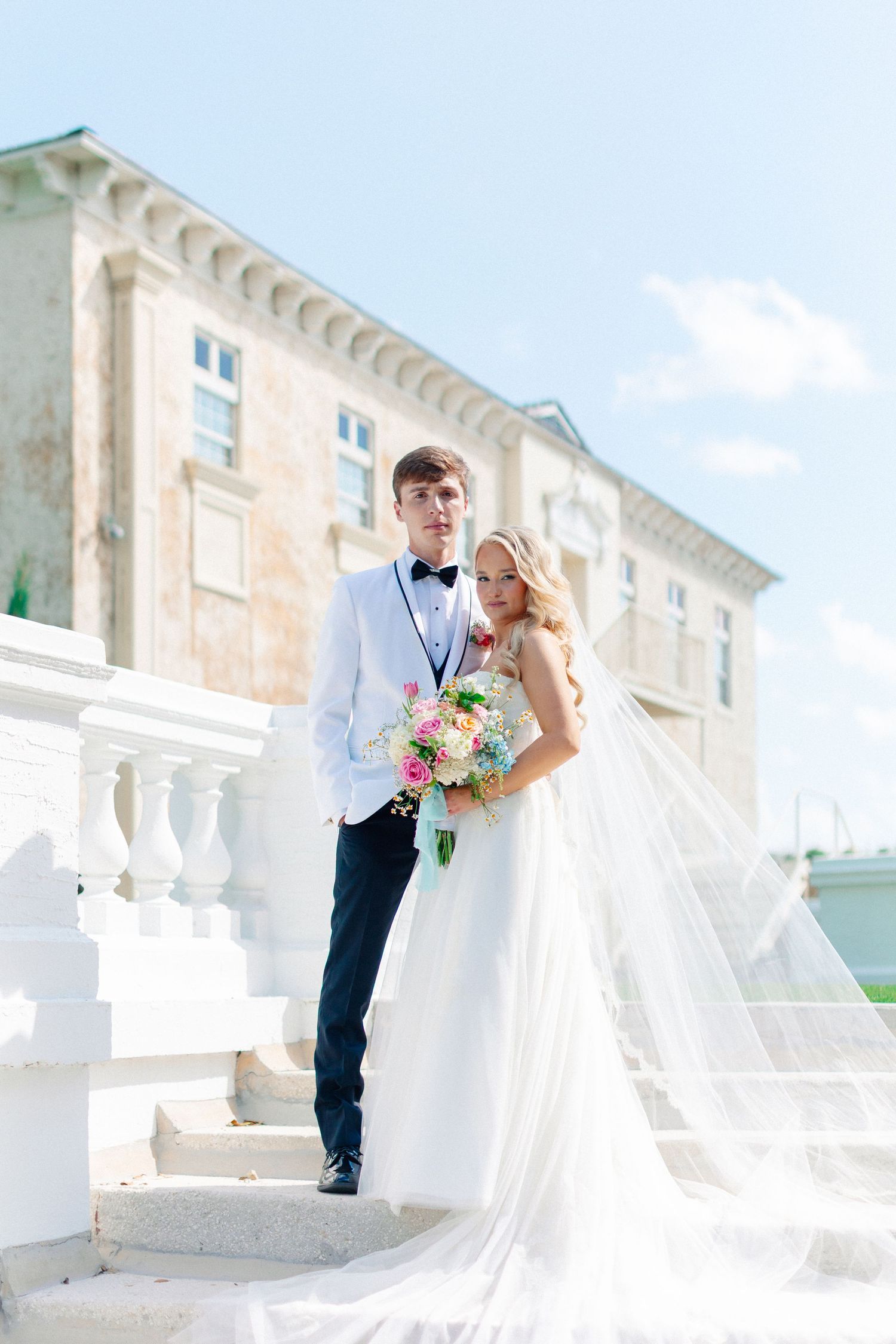 Elegant bride and groom posing on white marble steps outside a grand venue on a sunny wedding day, wedding at Bella Cosa