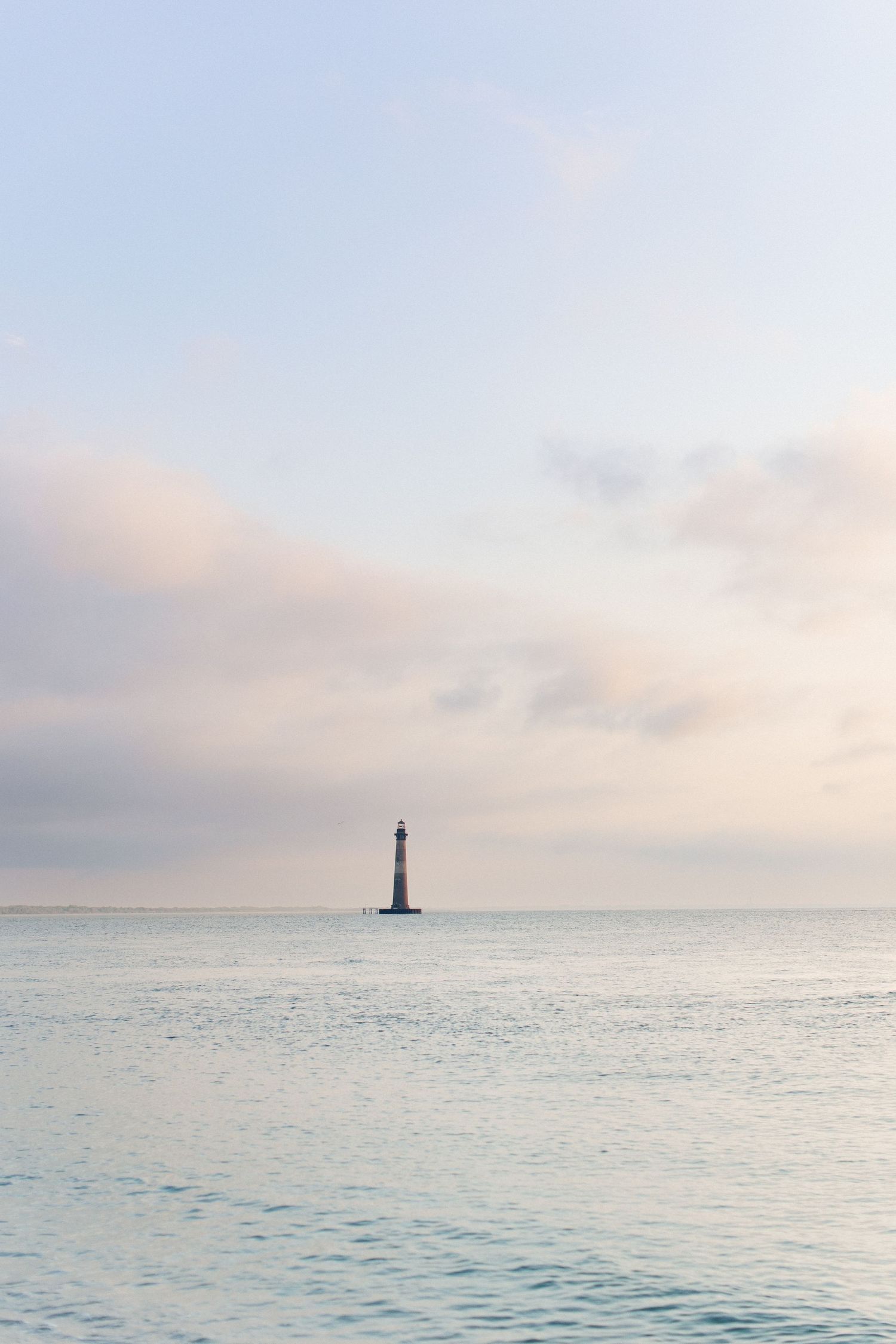 A solitary lighthouse stands in calm pastel ocean waters under a soft pink and blue cloudy sky at dusk.