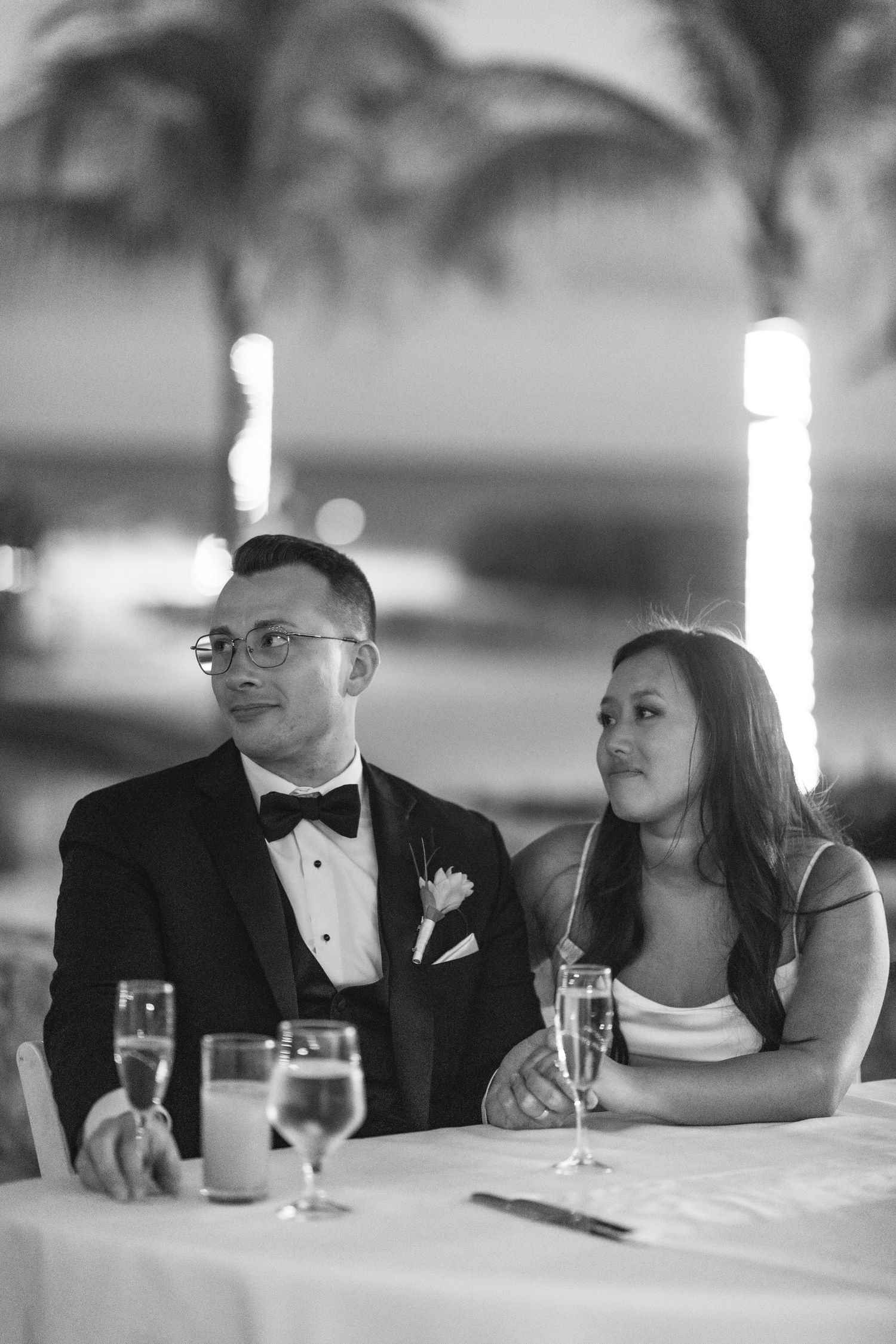 Black and white photo of a bride and groom sitting at a wedding reception table with champagne glasses, palm trees in background.