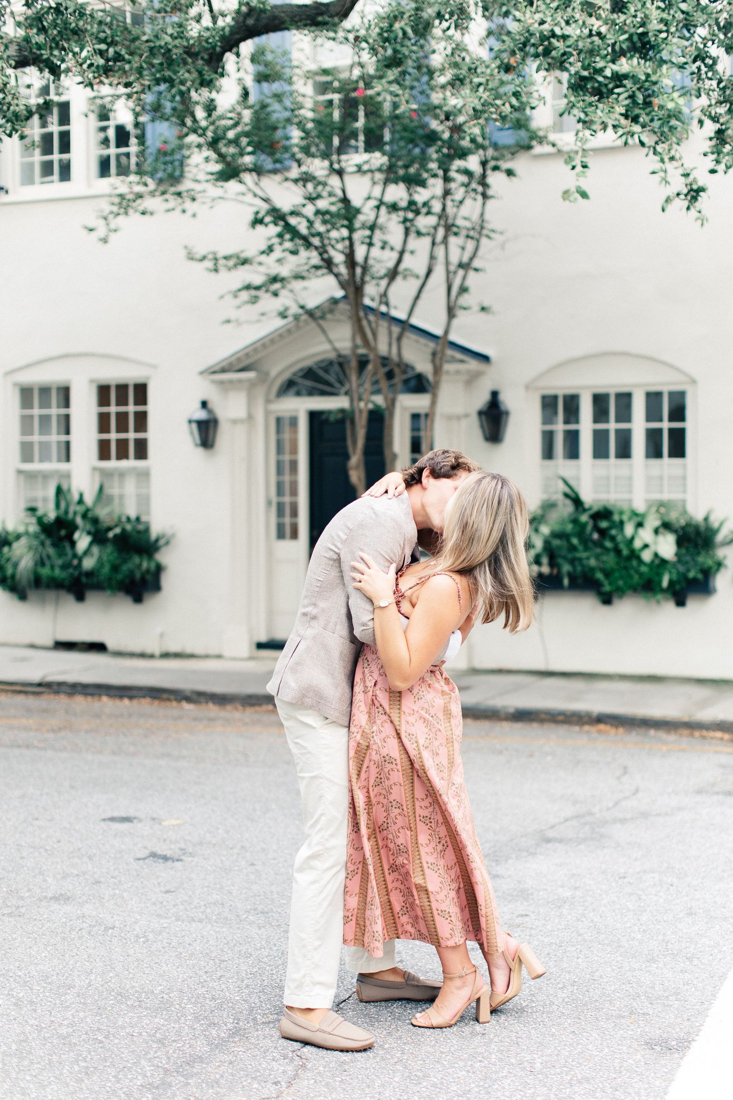 Couple sharing a romantic kiss on a street in front of a white building with lush greenery.