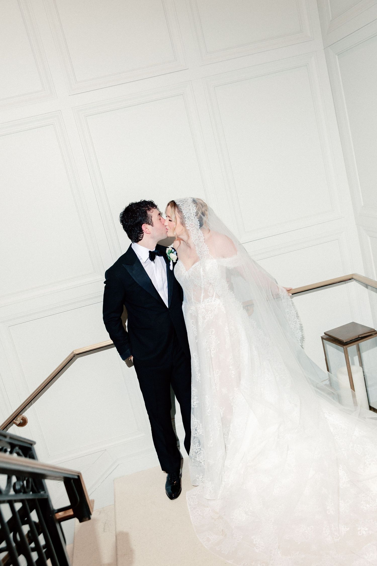 Bride and groom share a romantic kiss on an elegant staircase, bride in lace gown with veil, groom in black tuxedo.