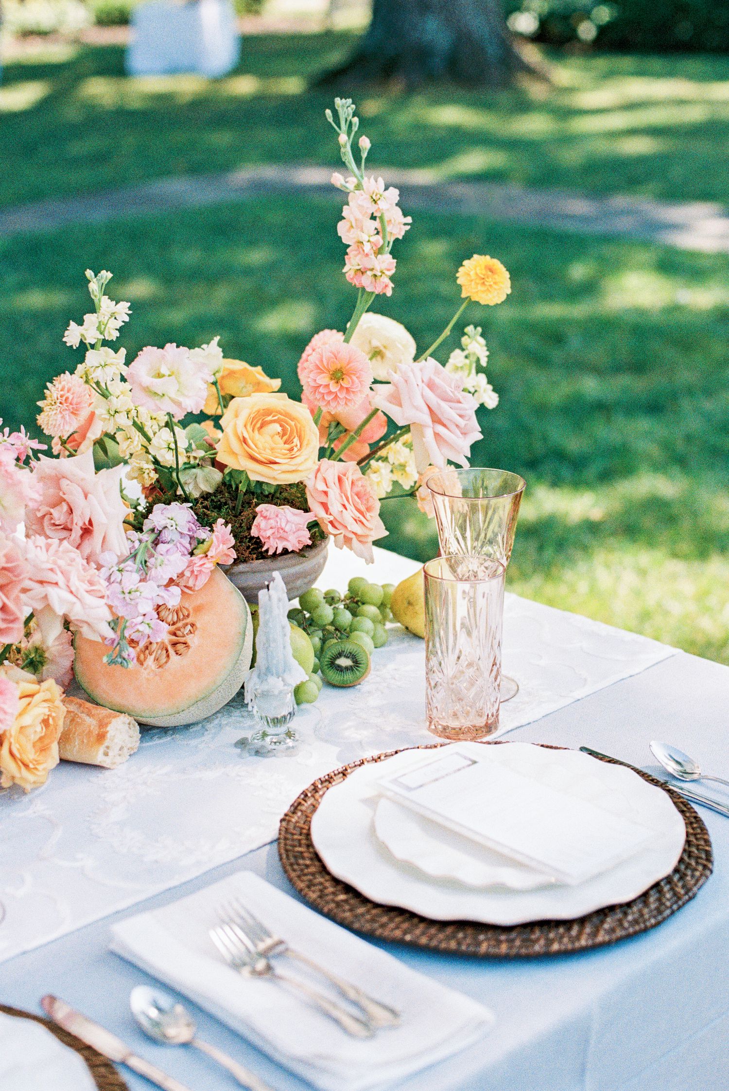 Romantic outdoor wedding tablescape with peach and pink floral arrangement, wicker charger plates, and fresh fruit accents.