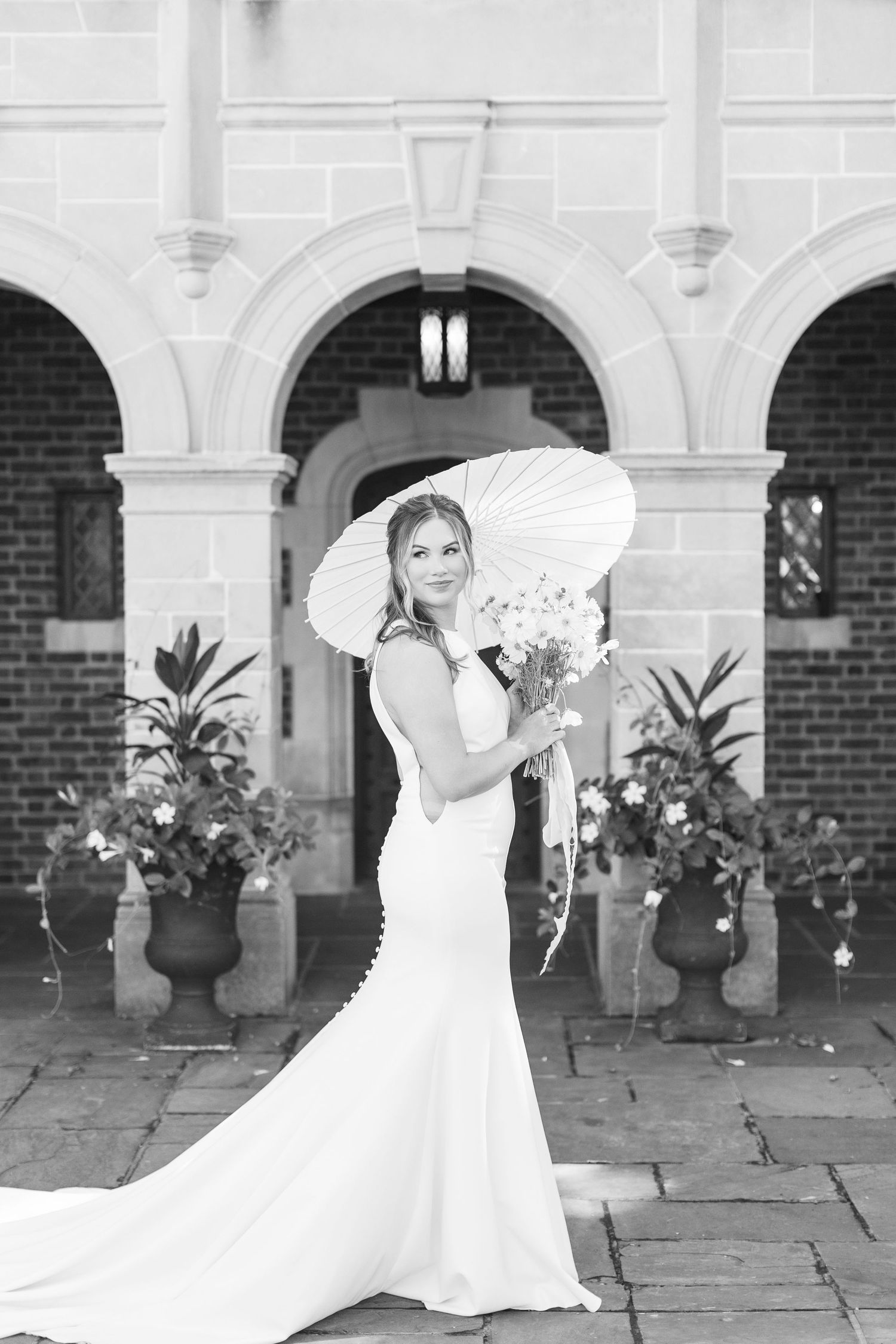 Bride in white gown holding parasol and bouquet poses before arched stone building entrance in black and white photo.