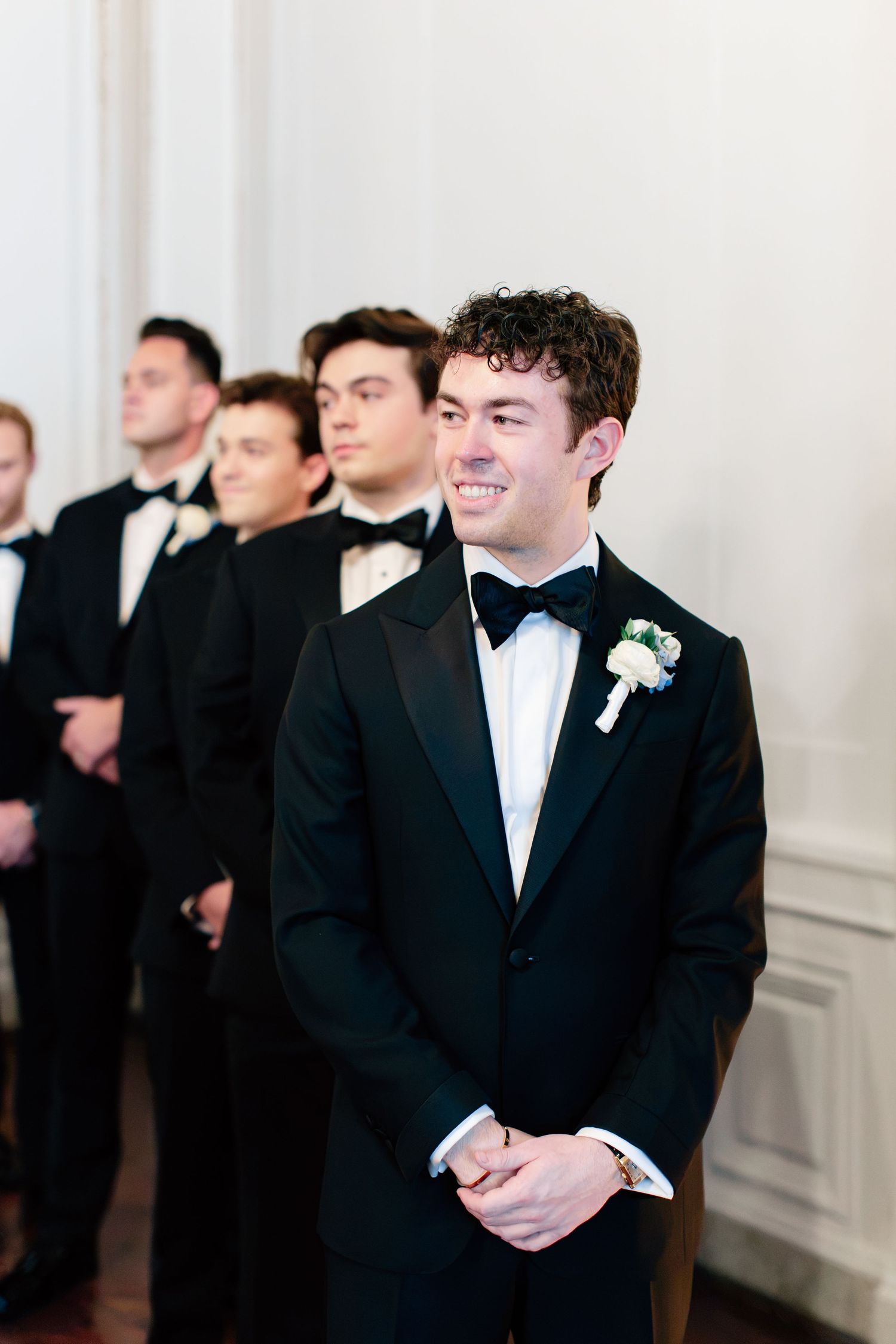 Smiling groom in black tuxedo with boutonniere stands at altar with groomsmen lined up behind him at wedding ceremony.