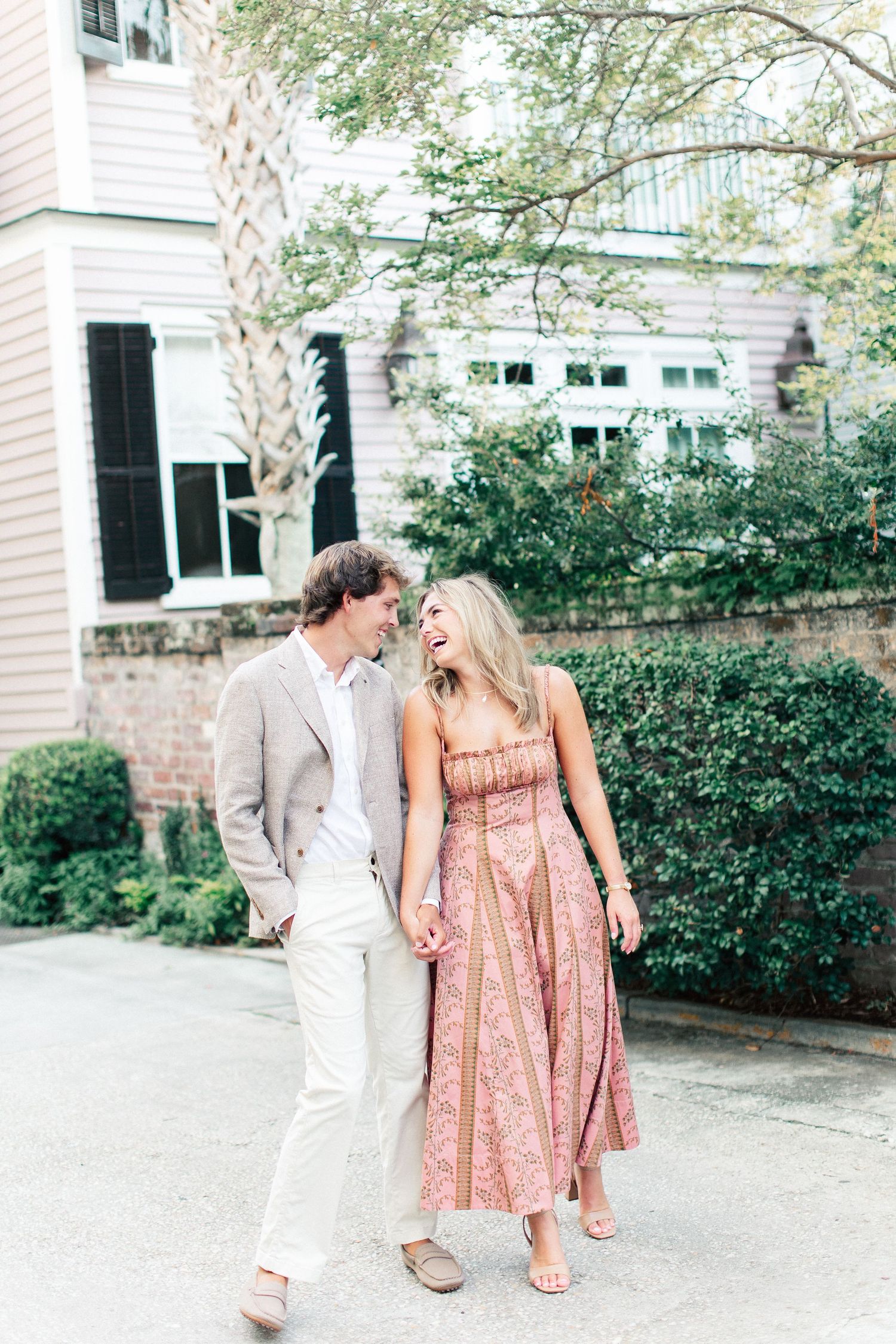 A laughing couple holds hands while strolling past a historic home surrounded by lush greenery.