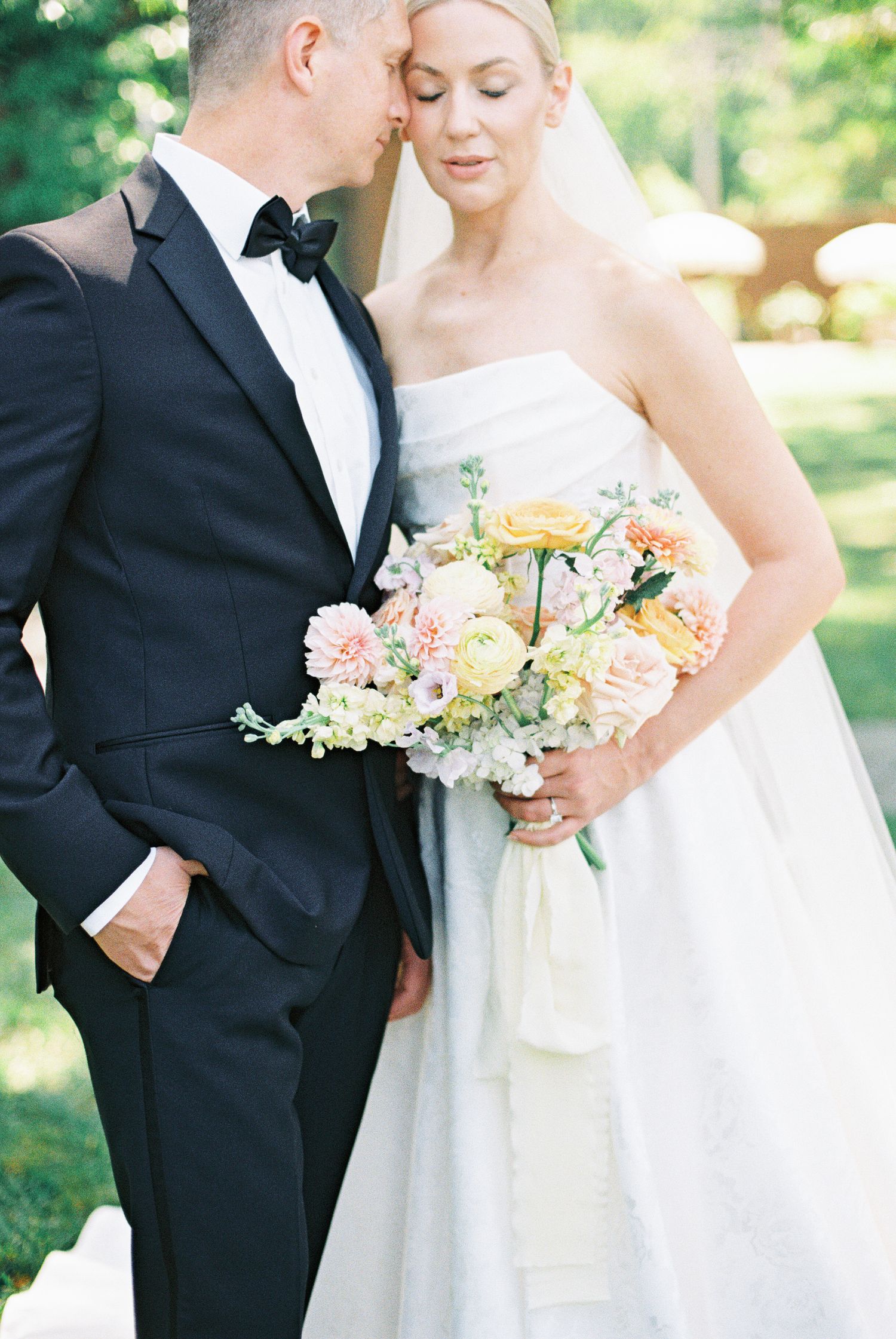 Groom in black tuxedo leans close to blonde bride holding colorful peach and pink bouquet in garden setting.