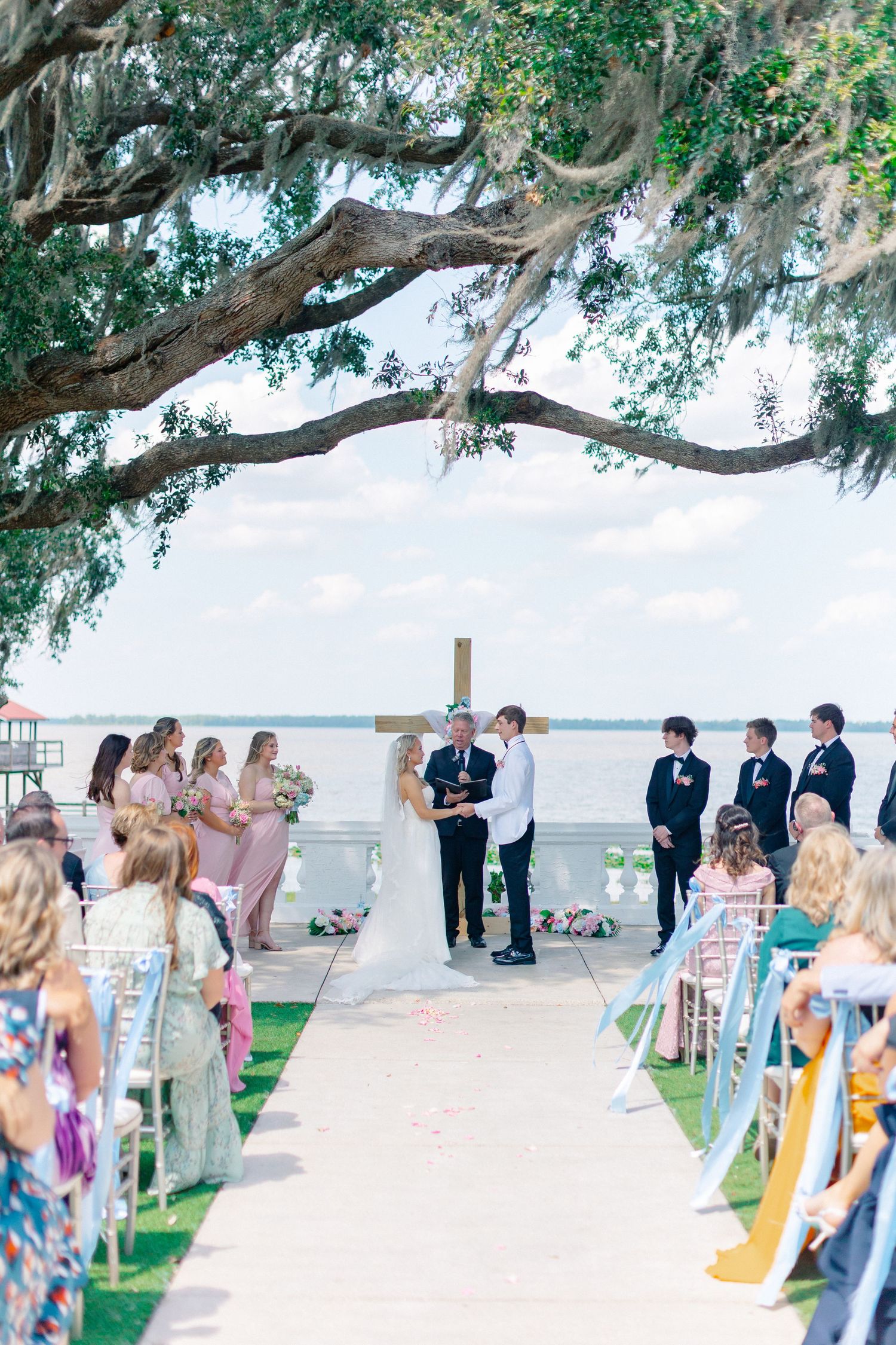 Outdoor waterfront wedding ceremony under a sprawling oak tree with bridesmaids in pink and groomsmen in black suits, Florida