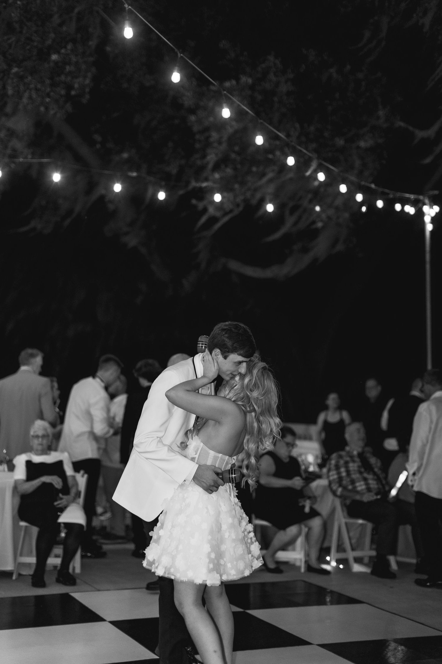 Couple shares first dance under string lights at outdoor wedding reception on black and white checkered dance floor, wedding at Bella Cosa