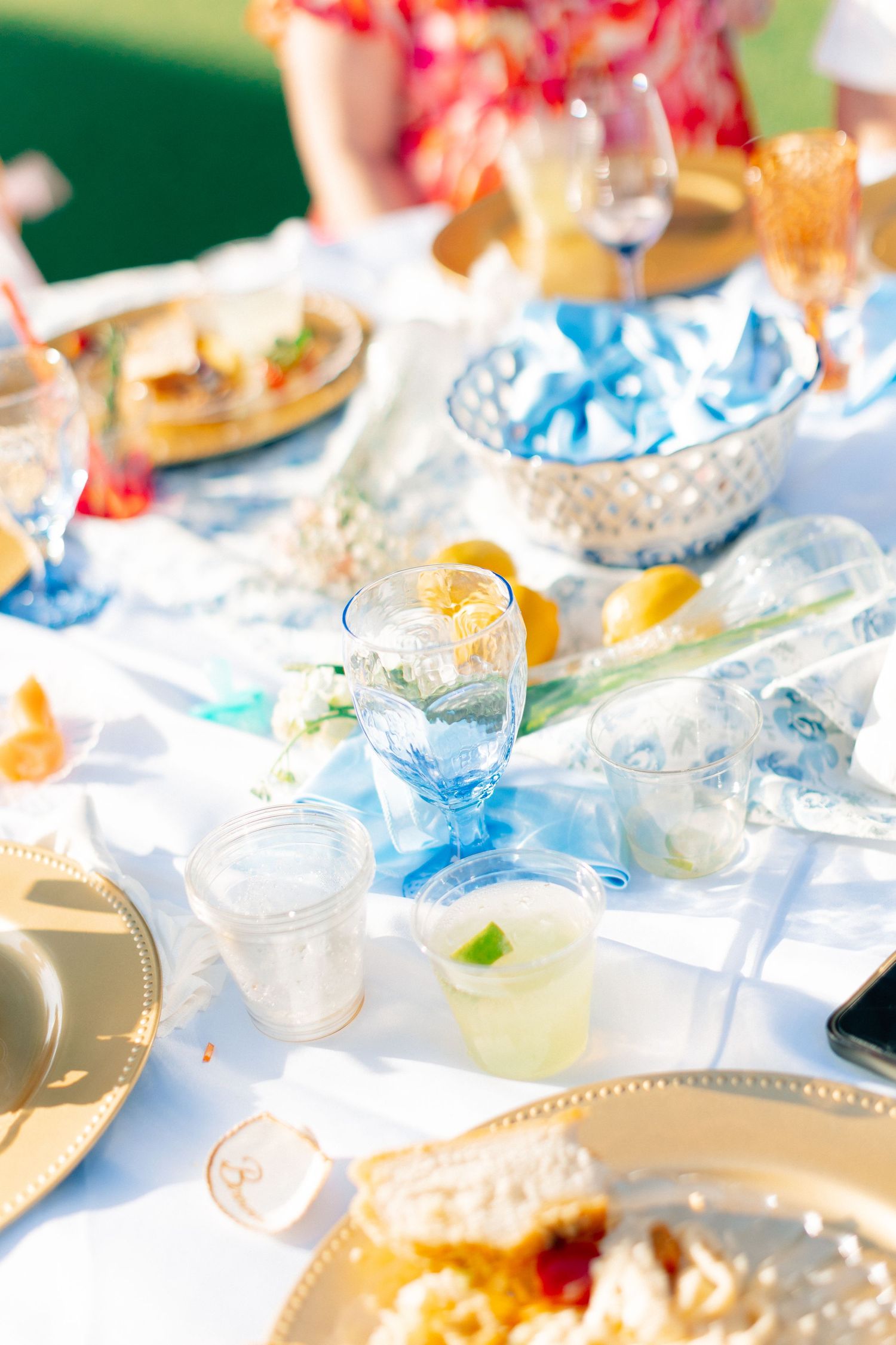 Elegant outdoor party table with blue glassware, gold charger plates, lemonade, and fresh fruit on a white tablecloth, wedding at Bella Cosa