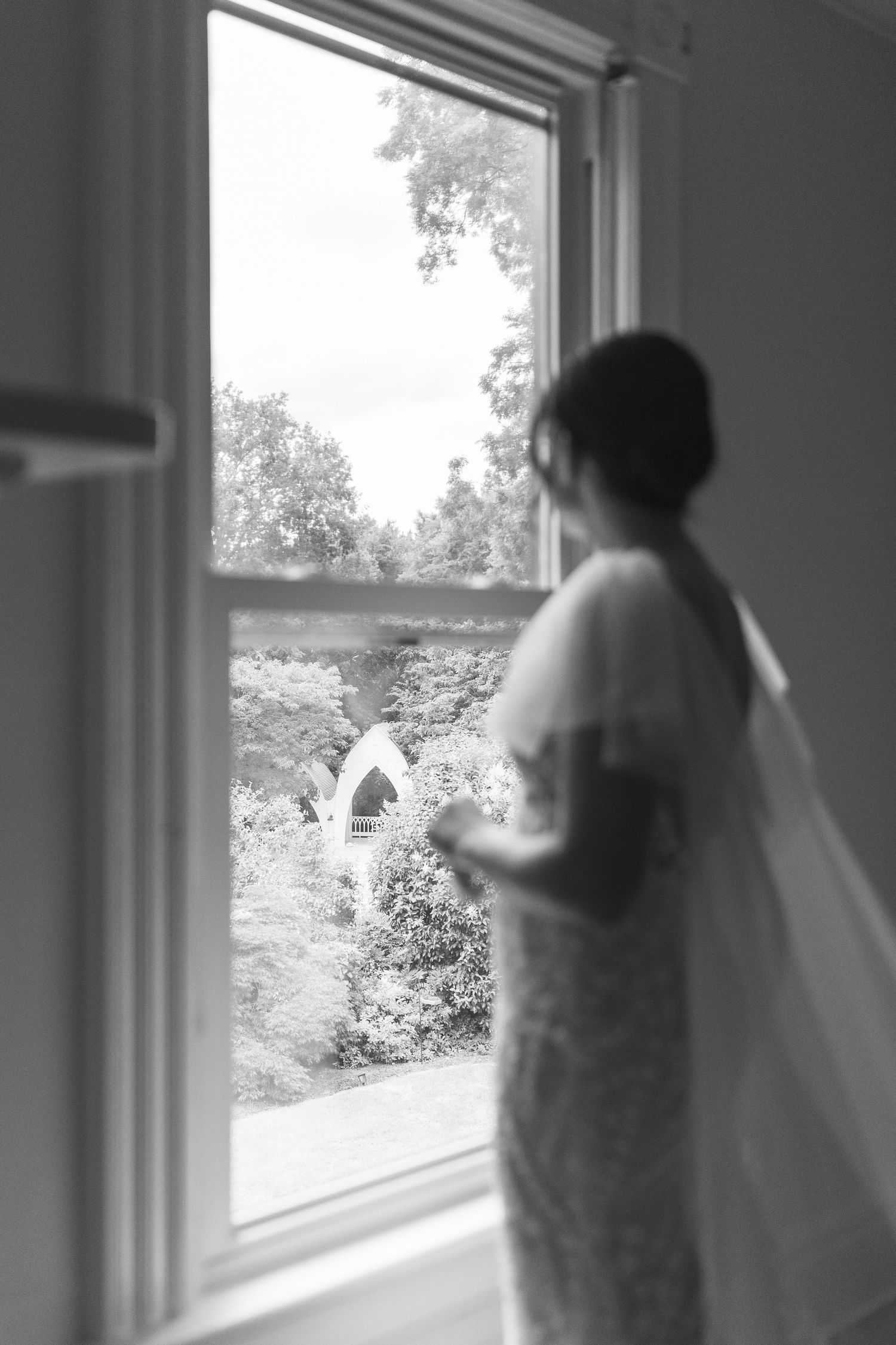 Black and white photo of a bride in a veil gazing out a window at a garden wedding arch.