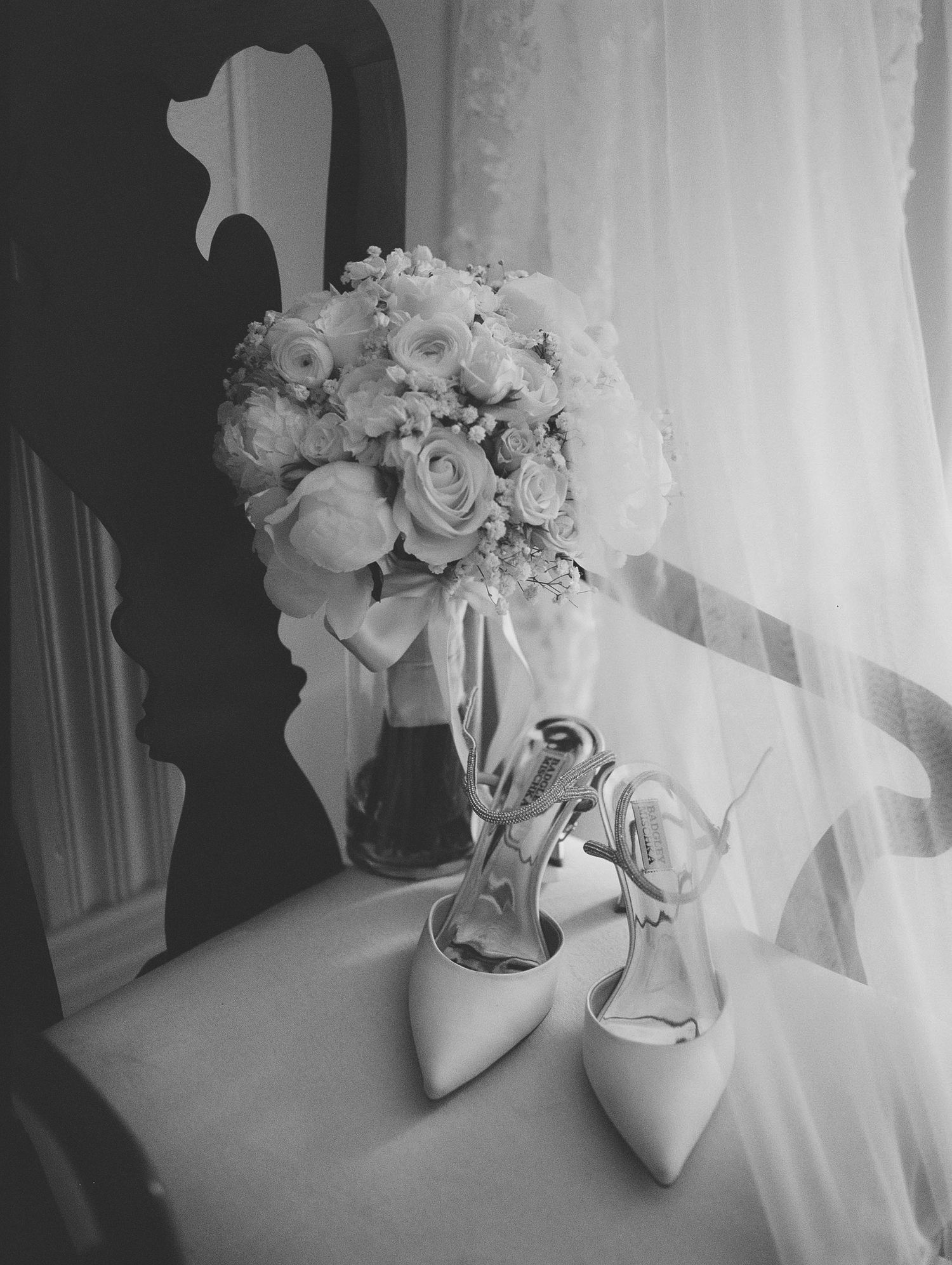 Black and white wedding photo of bridal bouquet with roses and elegant heels on a table near a window.