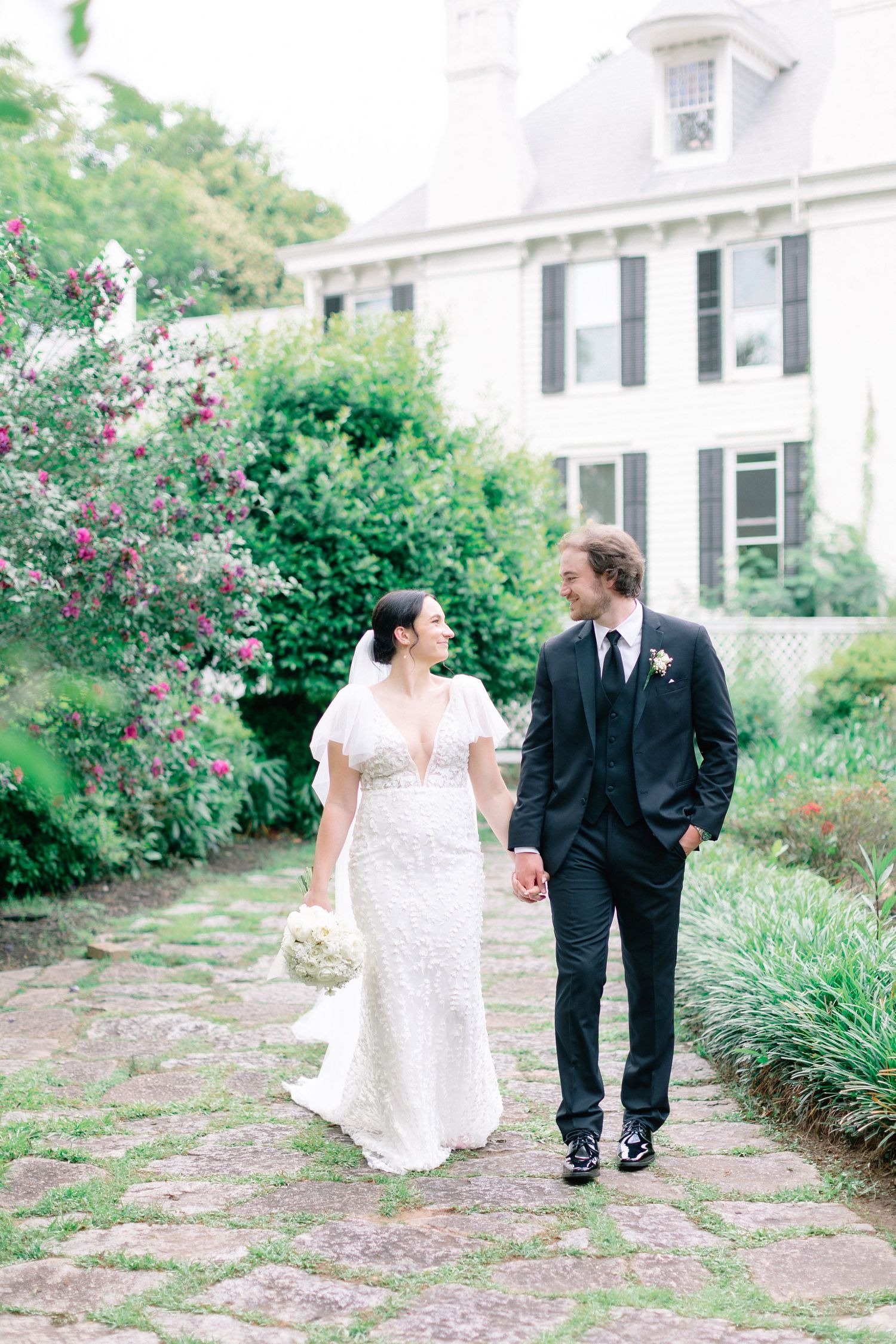 Bride and groom walk hand-in-hand along a stone path in a lush garden beside a white colonial mansion.