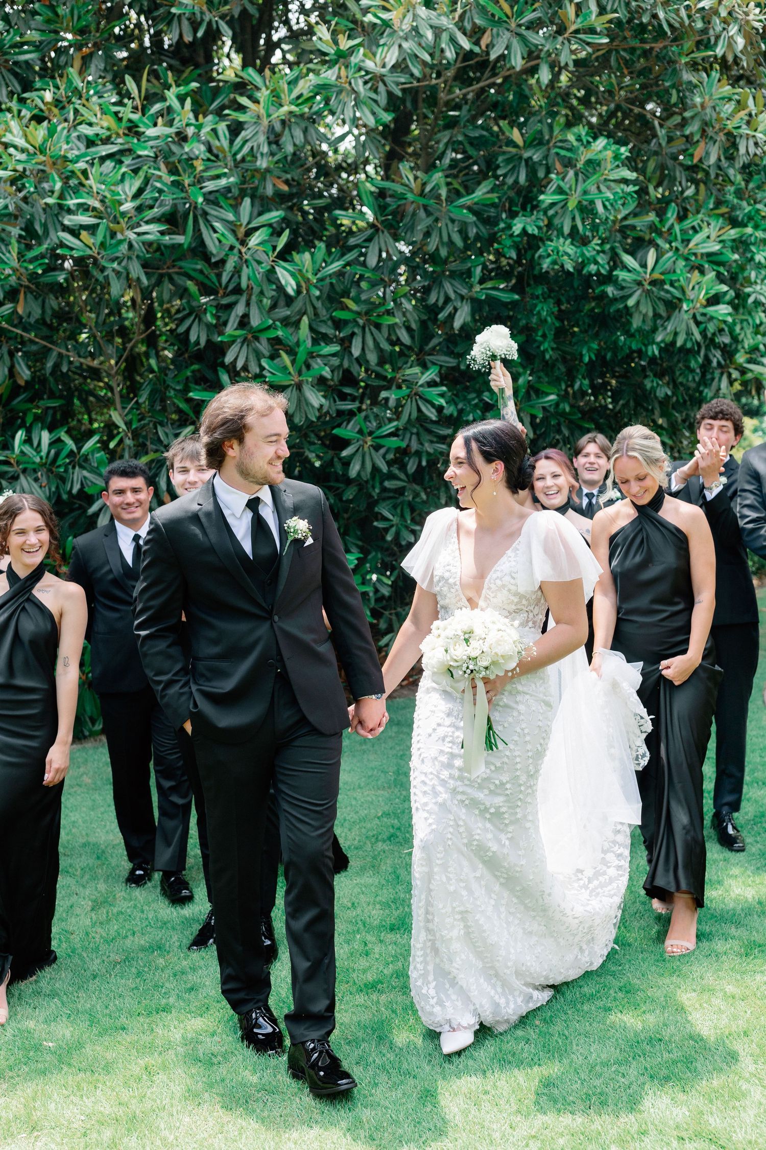 Bride and groom hold hands walking with wedding party in black attire on green lawn before lush trees.