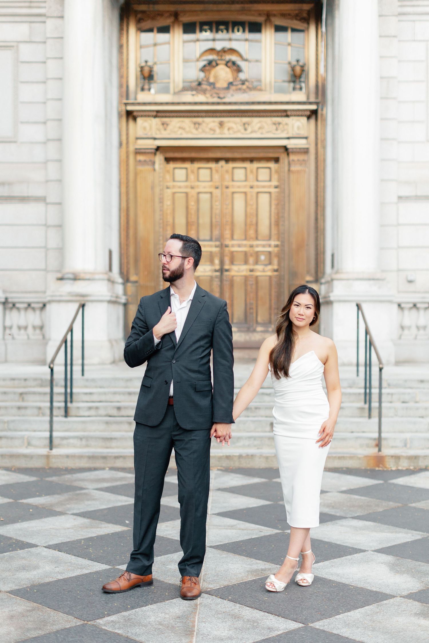 Couple posing outside ornate building, man in dark suit with glasses, woman in white strapless midi dress holding hands.