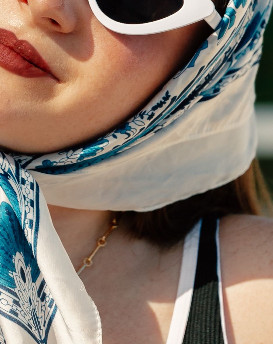 Woman wearing white sunglasses and a blue floral silk head scarf with a gold chain necklace.