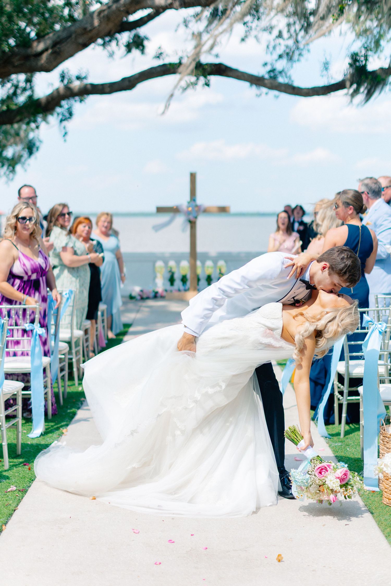 Groom dips bride for a kiss at outdoor waterfront wedding ceremony with guests seated along the aisle.